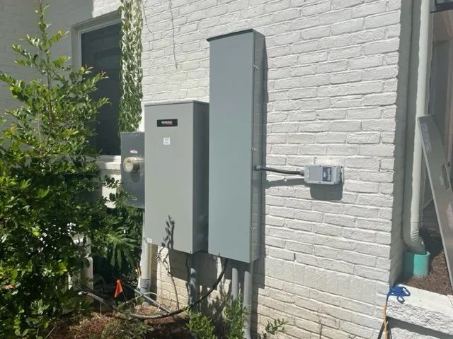 Electrical utility boxes mounted on exterior white brick wall with a small window, surrounded by green plants, pipes, and a utility blue cap with a cable.