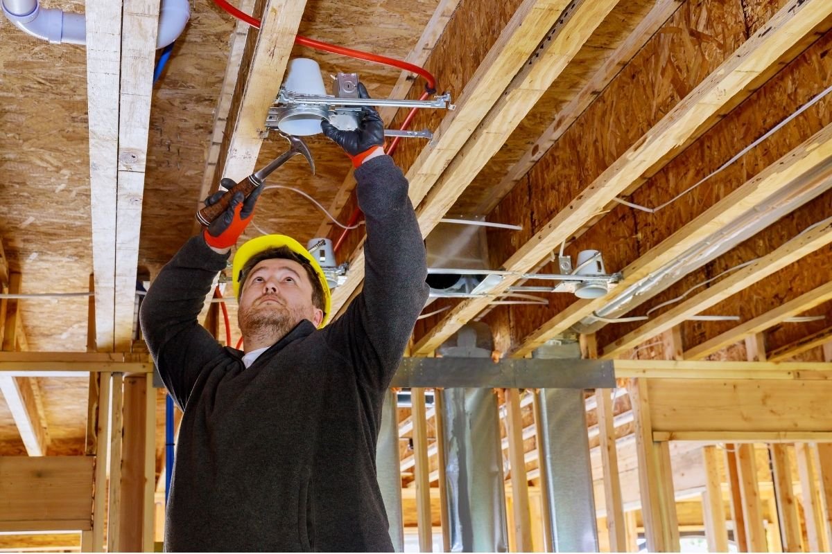 A construction worker wearing a yellow safety helmet, black gloves, and a dark sweater installs ductwork on a building's ceiling. The space has exposed wooden beams and construction materials.