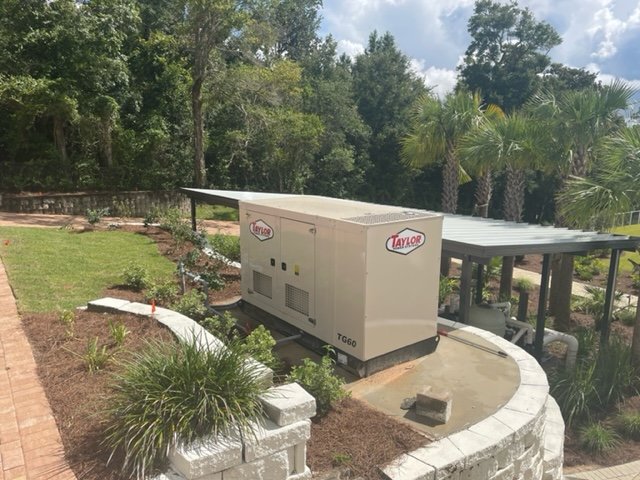 A Taylor generator is set up on a concrete pad outdoors, surrounded by landscaped garden beds with plants, bordered by a white brick wall, and shaded by trees and palm trees in the background.