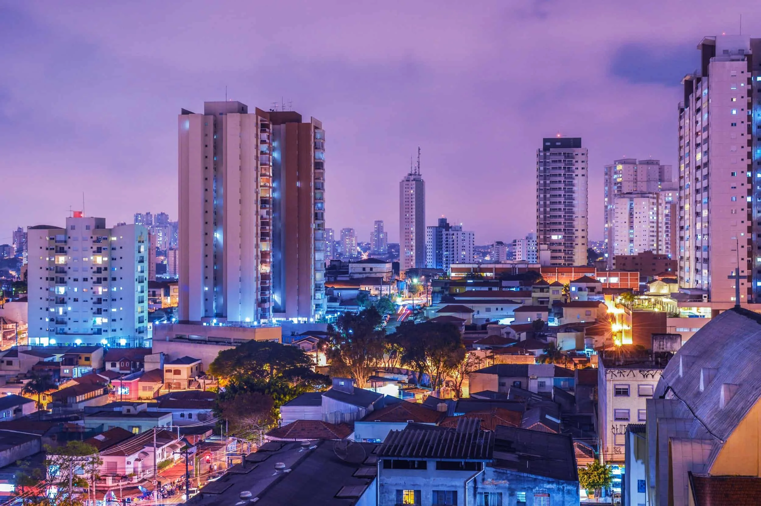 Night view of a city skyline with tall buildings illuminated by blue and purple lights, with lower residential buildings and trees in the foreground.