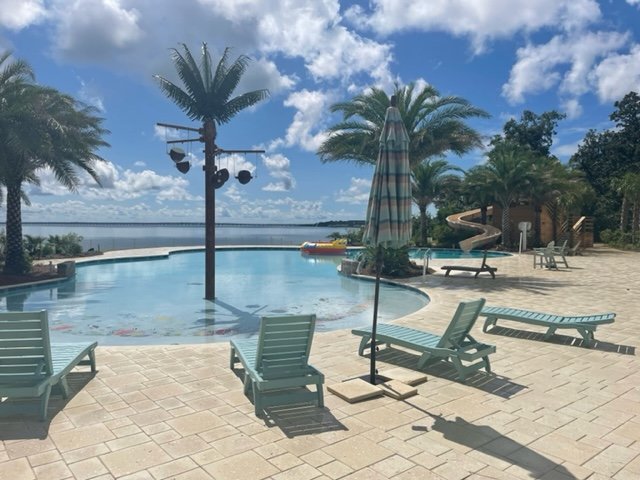 Empty swimming pool area with lounge chairs, umbrellas, palm trees, and a water slide, under a partly cloudy sky.