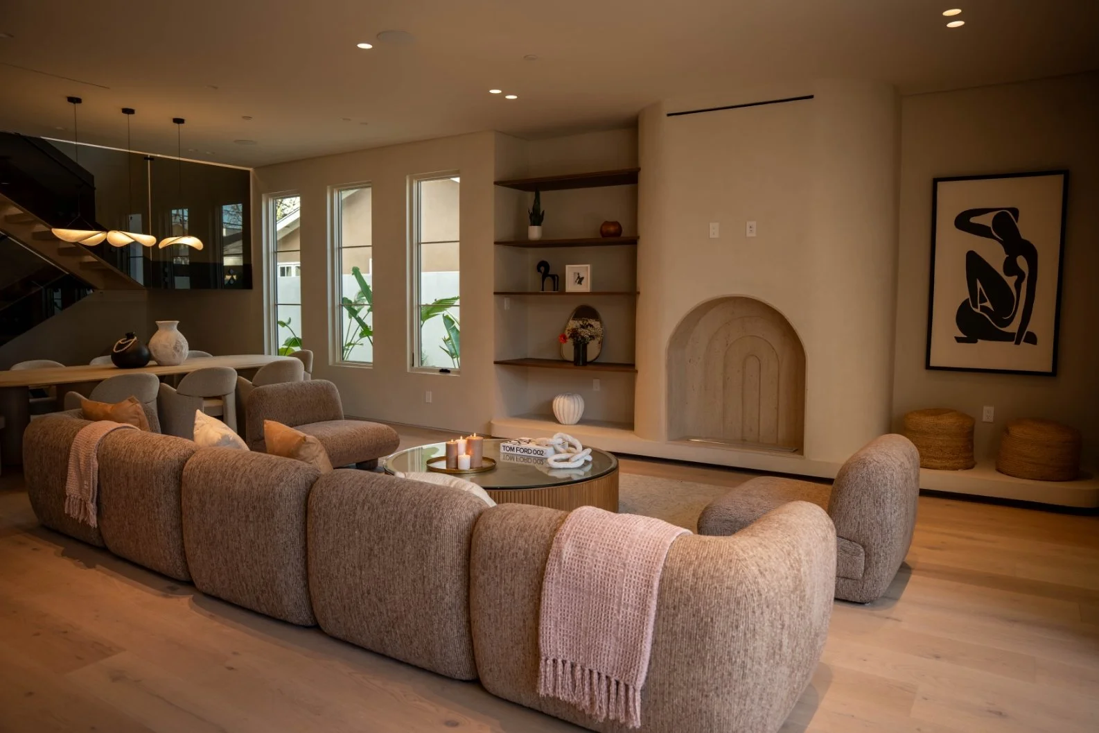 A modern living room with a large beige sectional sofa, a round coffee table with candles, a built-in wall shelf with decorative items, a large framed abstract black and white art piece, and large windows with plants outside.