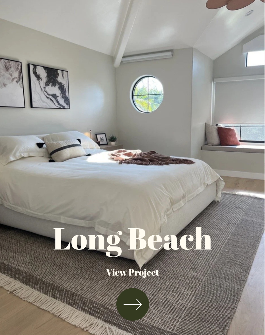 A bedroom with a large bed, white bedding, decorative pillows, and artwork on the wall. There is a circular window and a window seat with pillows. The room features light-colored walls, a wood floor, and a textured area rug.