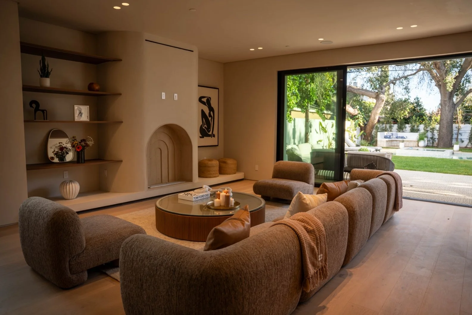 Living room with beige curved sectional sofa, glass-top round coffee table with candles, neutral wood flooring, large sliding glass door opening to a green backyard, built-in shelves with decorative items, and outdoor seating.