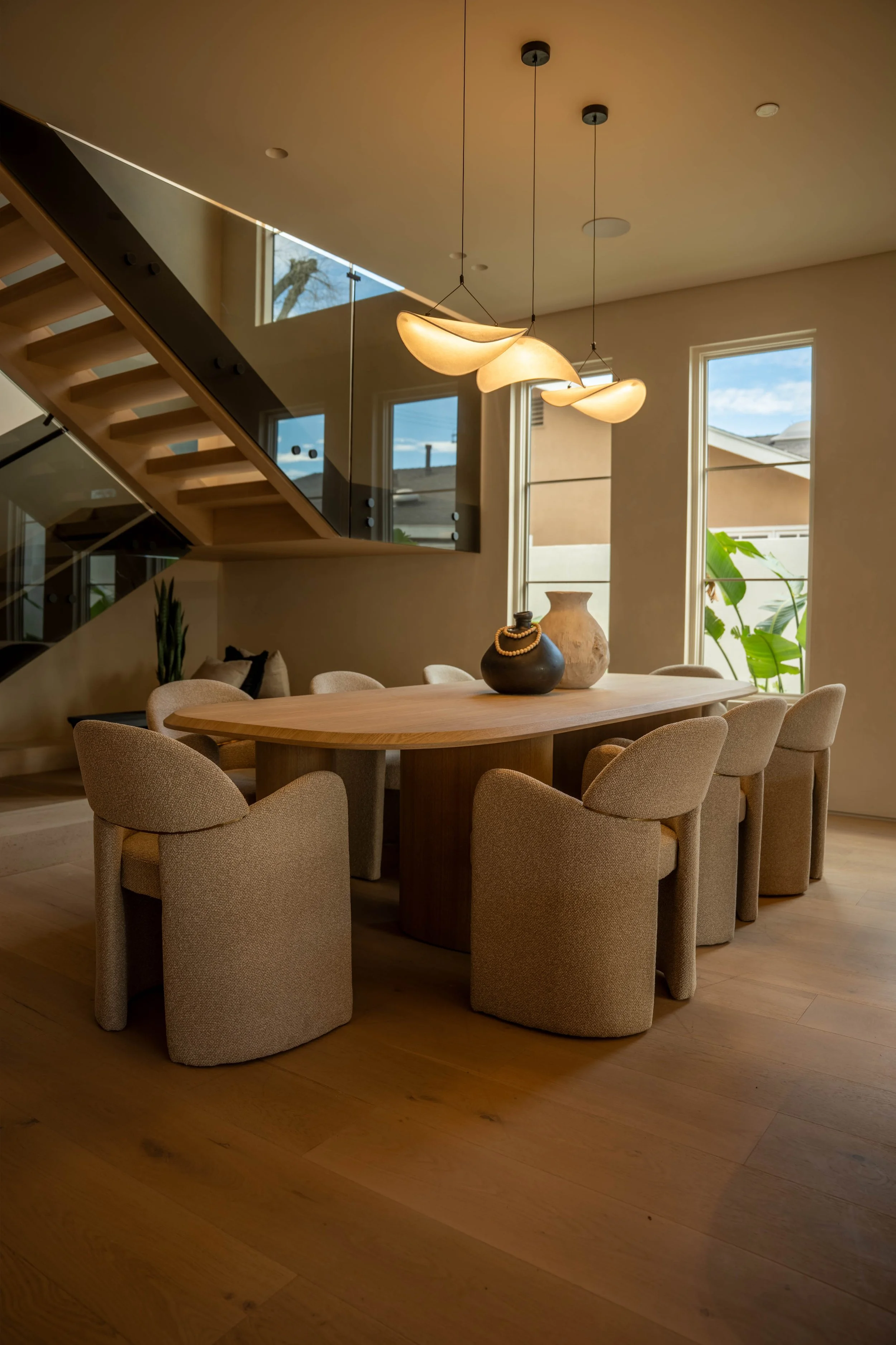 Modern dining room with a wooden table, beige upholstered chairs, and decorative vases. Large windows let in natural light, and a contemporary pendant light hangs from the ceiling.