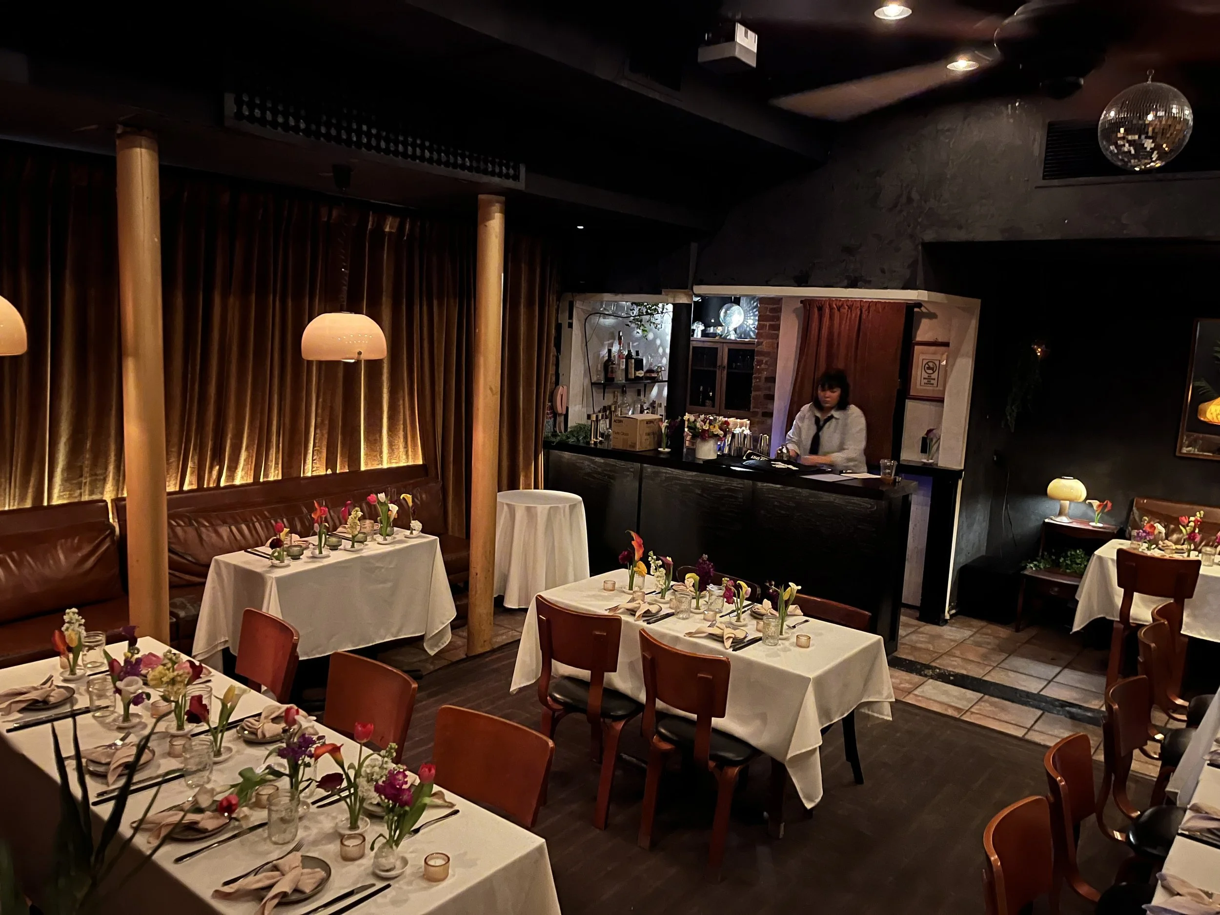Cozy restaurant dining area with tables set for a special occasion, decorated with colorful flowers and candles, soft dim lighting, dark wood furniture, and a visible bar in the background.
