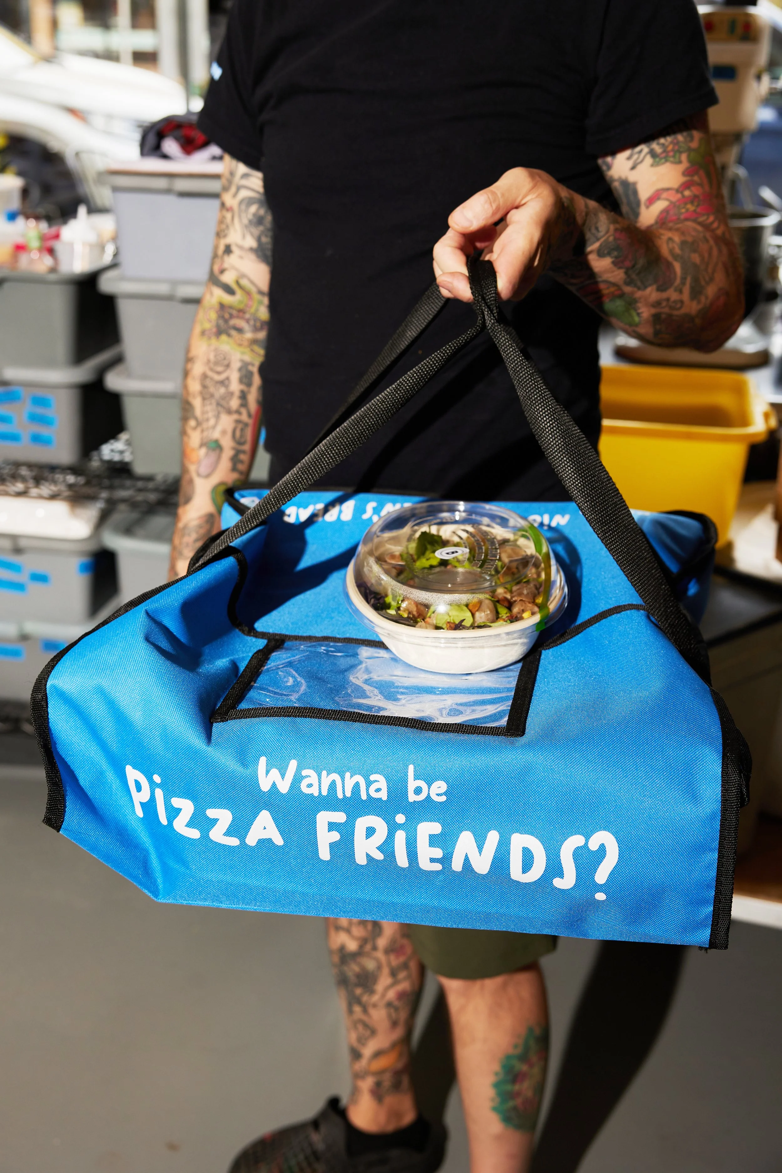 Man holding a blue pizza delivery bag with "Wanna be pizza friends" on the top, and a salad container balancing on top.