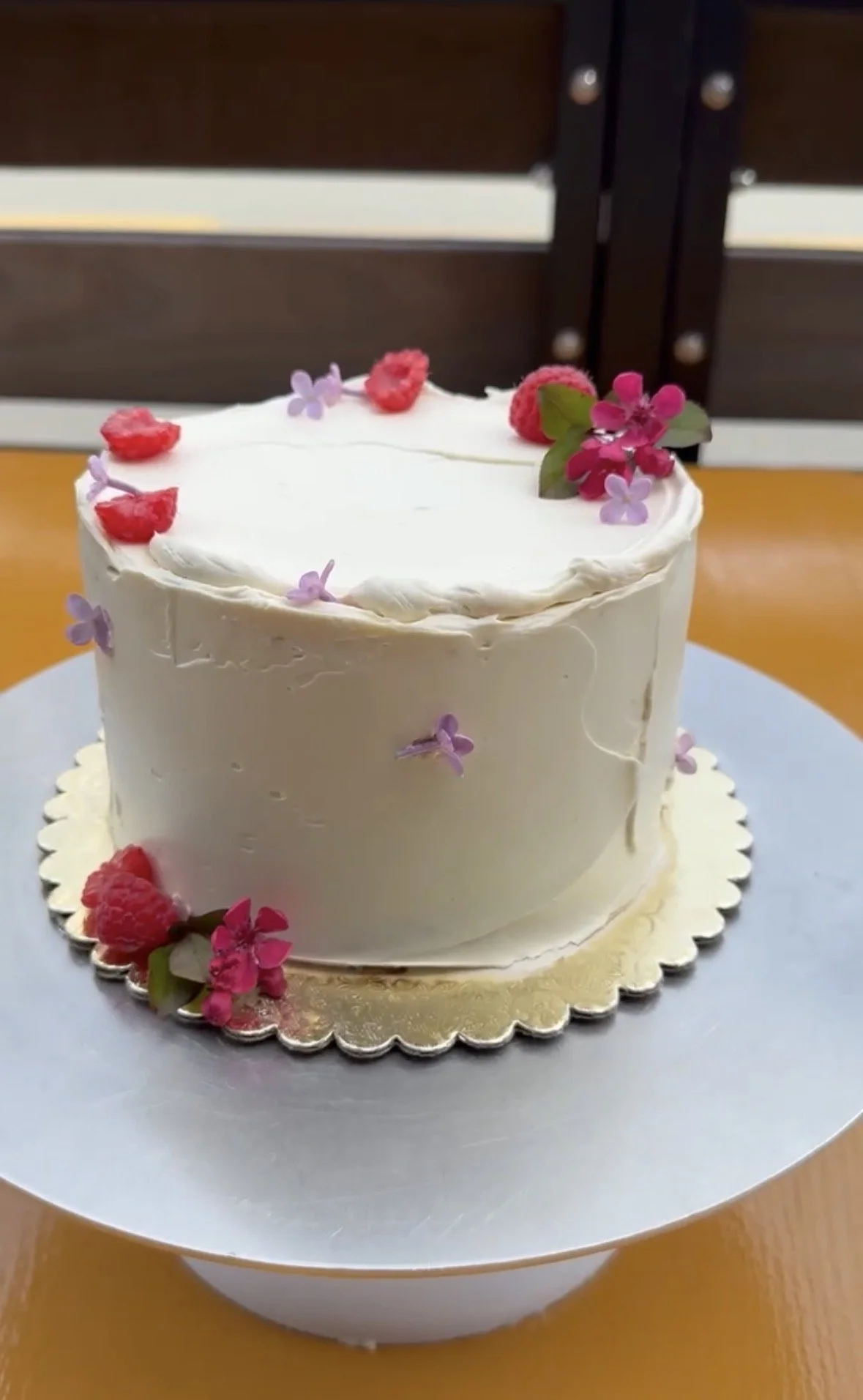 Vanilla cake adorned with edible florals and raspberries, on a gold scalloped rimmed pastry plate.
