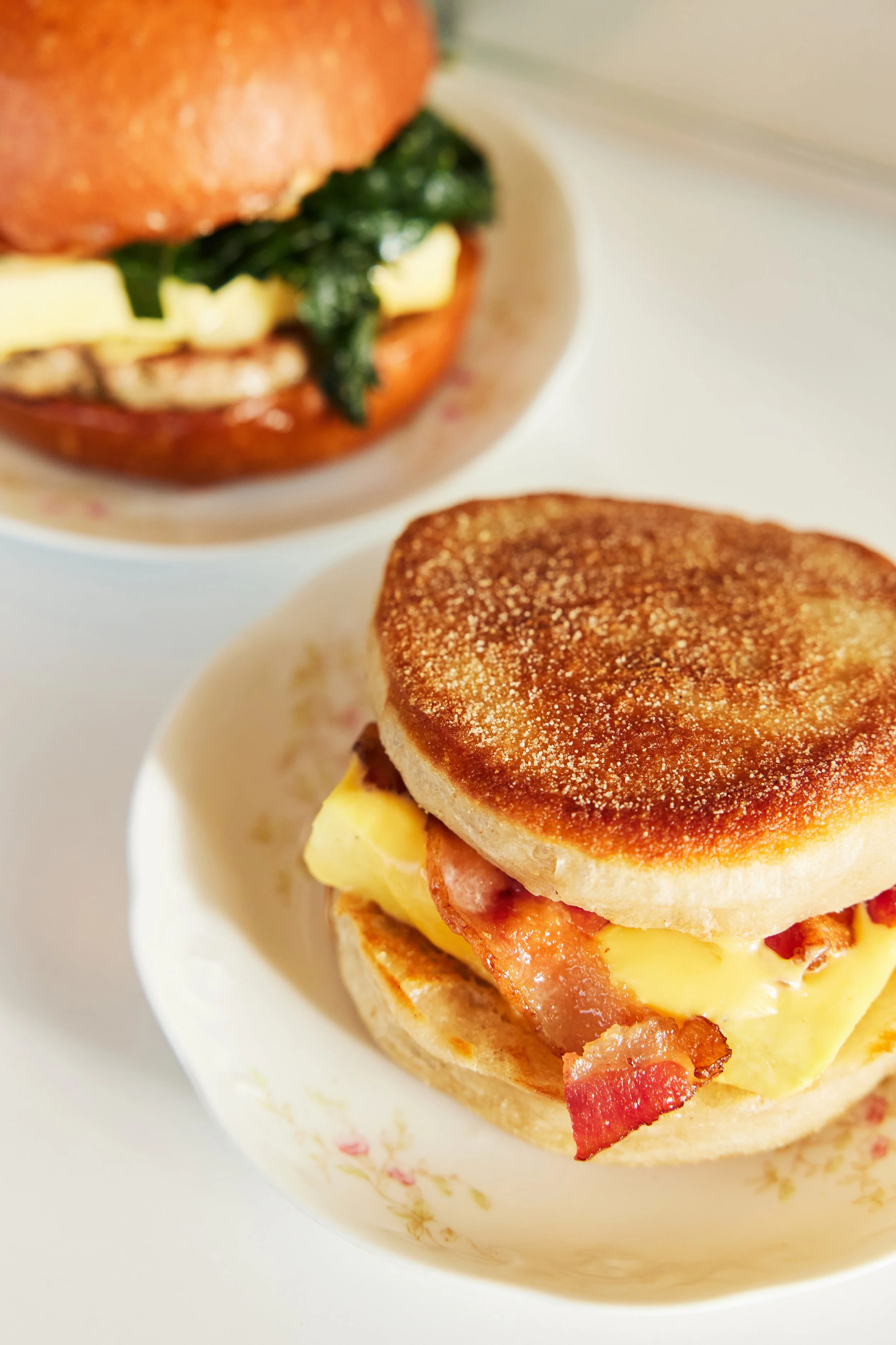 English muffin breakfast sandwich with bacon sitting on a plate in the foreground, with a brioche breakfast sandwich in the background.  As featured in Seattle Met Magazine.