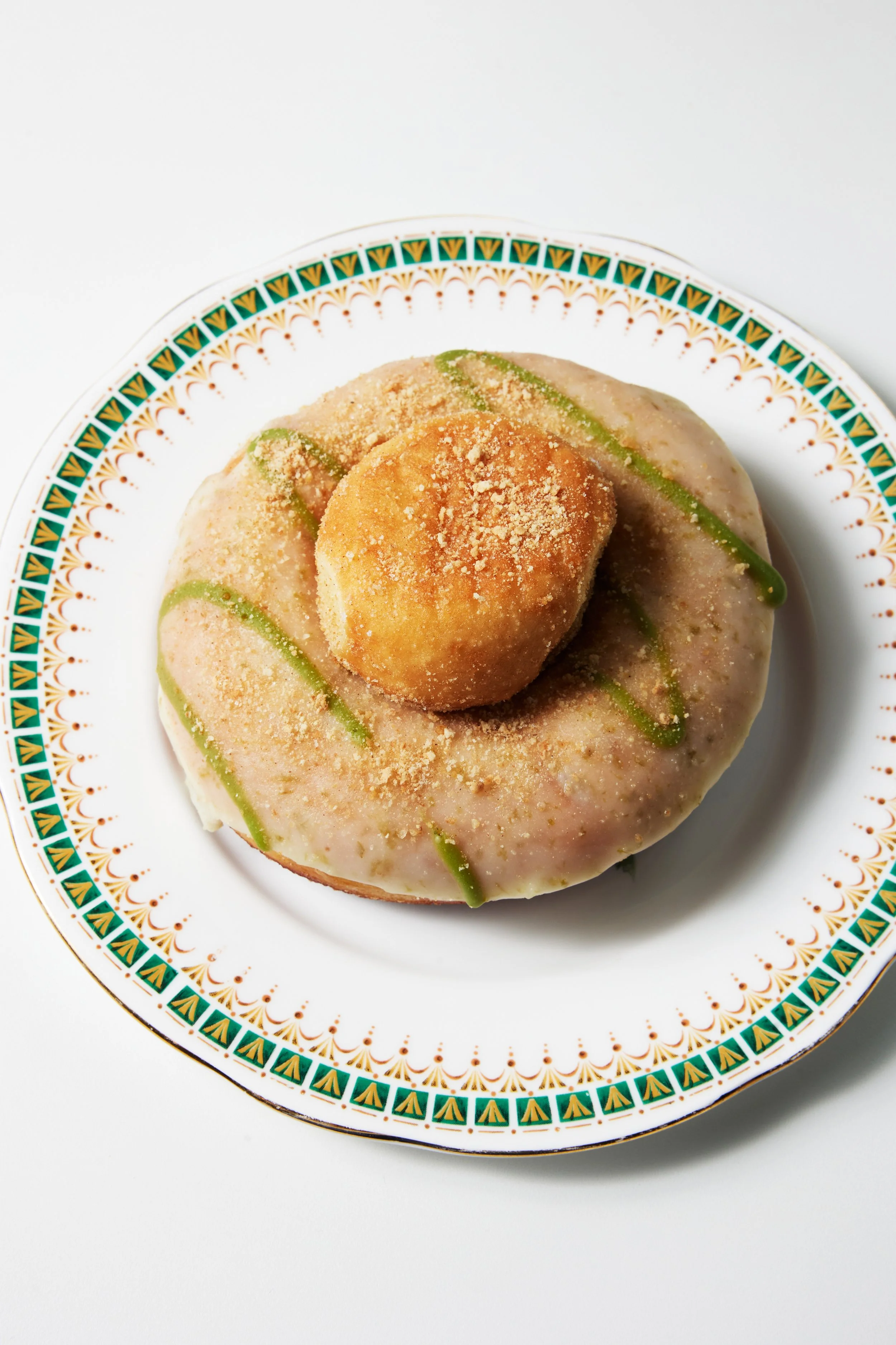 A donut with green matcha glaze topped with a donut hole, sitting on a white plate with green and yellow border
