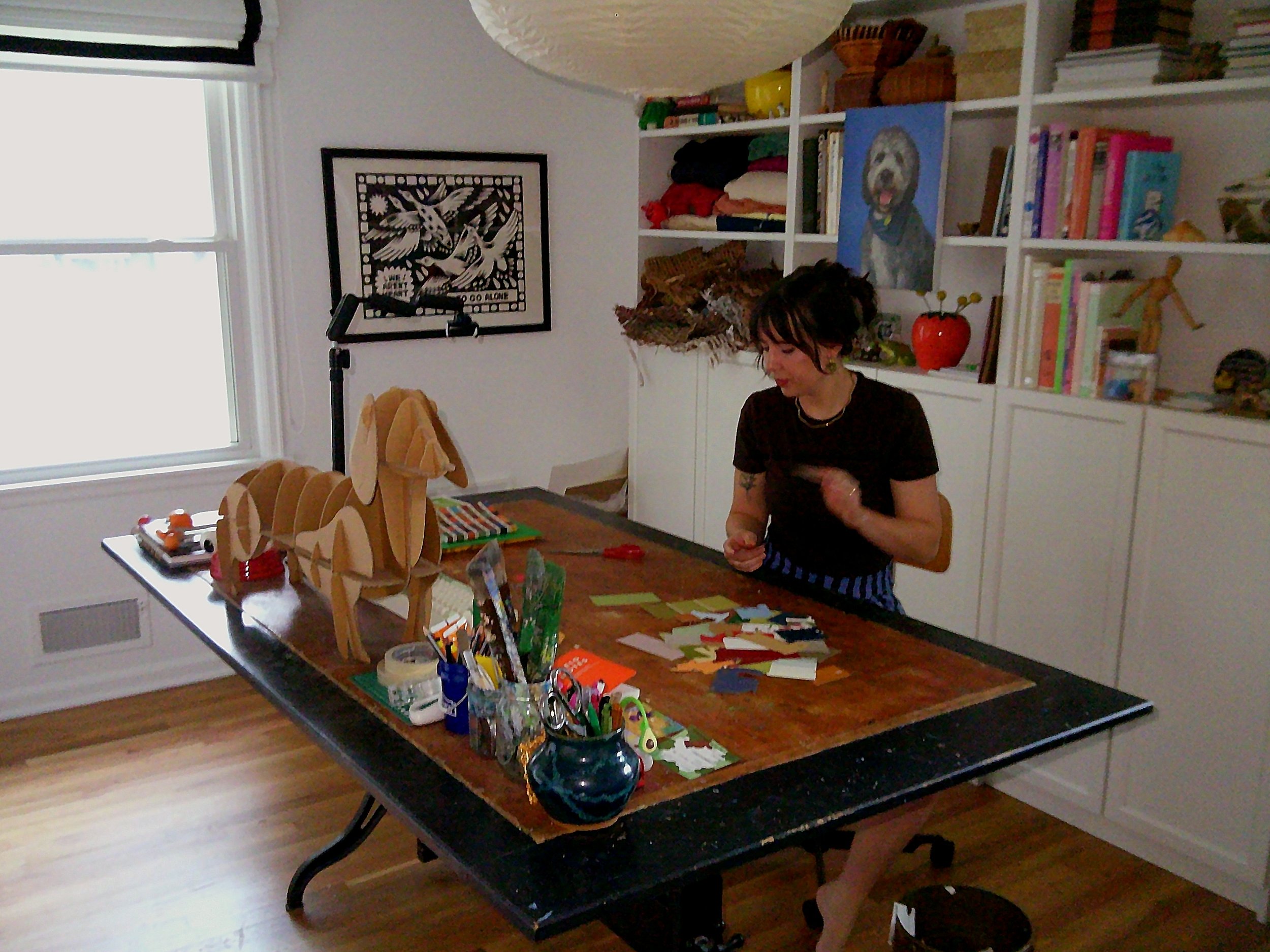 A woman sitting at a table with arts and crafts supplies, including paper, scissors, markers, and a wooden dog figure, inside a room with white bookshelf, artwork, and a window.