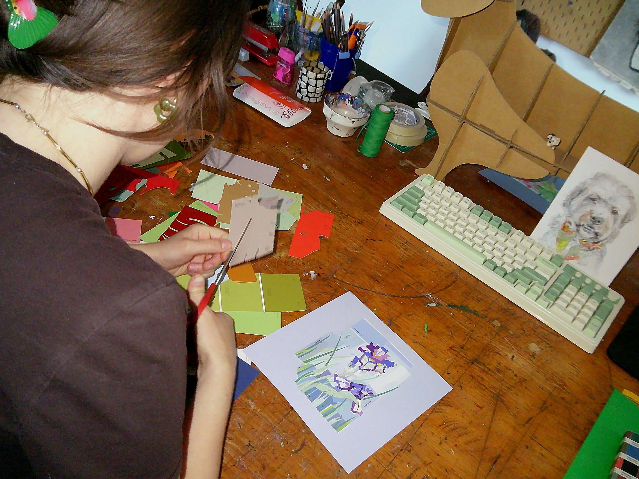 A person in a brown shirt with earrings is sitting at a wooden table, engaging in arts and crafts. The table is cluttered with colorful paper, scissors, and other craft supplies. In the background, there are organized containers with pens, scissors, and rolls of tape, along with a keyboard and a greeting card with a dog illustration.
