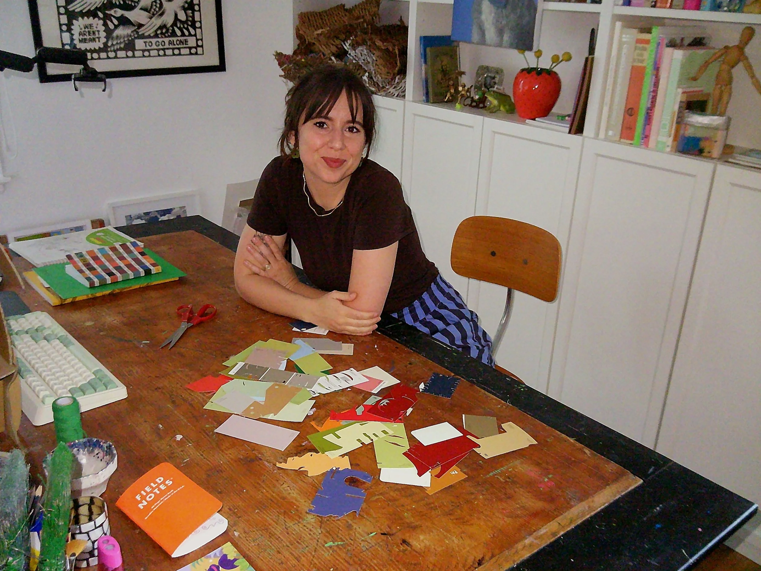 A woman sitting at a crafting table with various colored papers, scissors, and crafting supplies. Shelves with books and decorative items in the background.