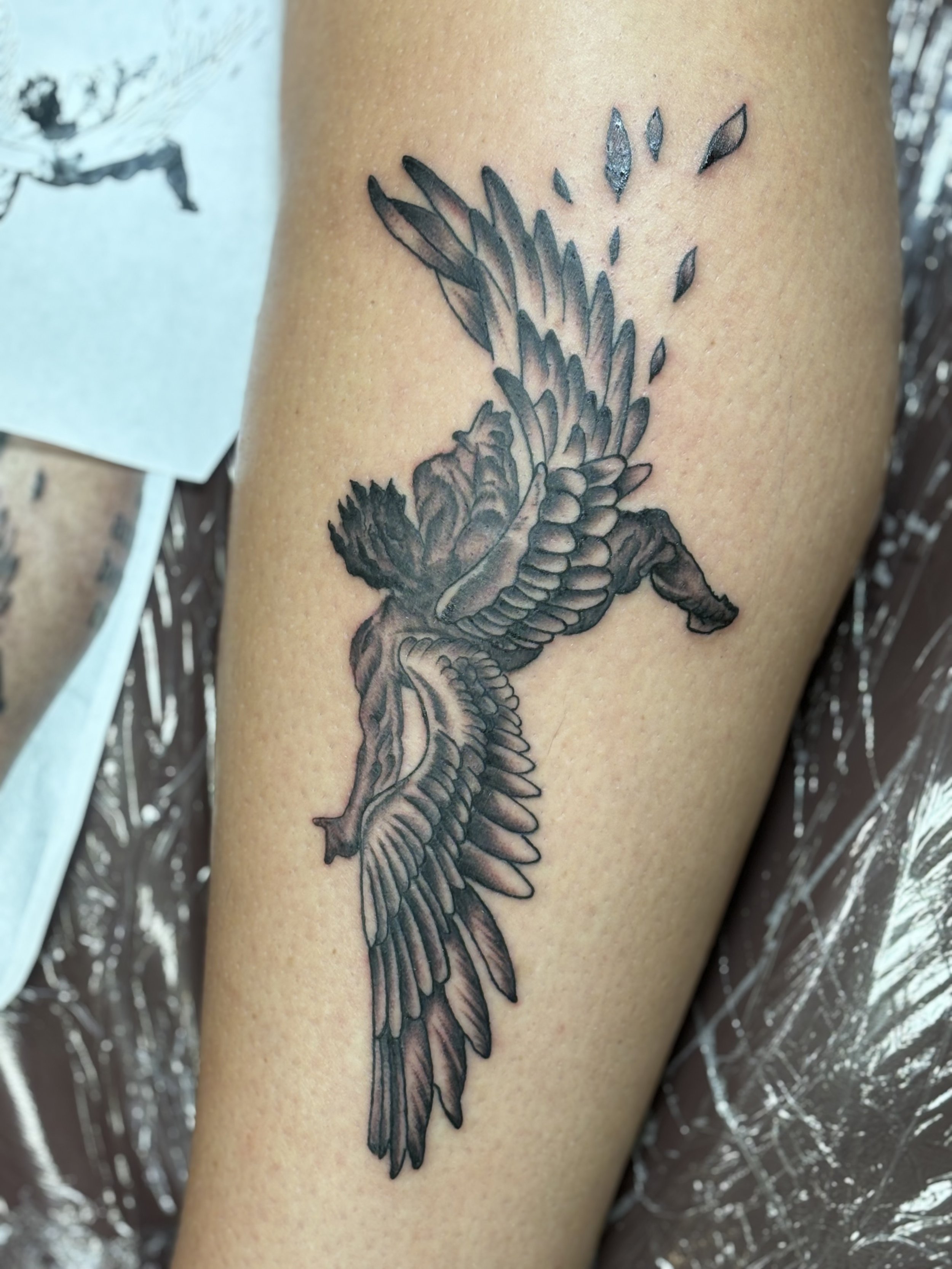 Tattoo of angel in grayscale with wings outstretched on arm, adjacent to a tattoo reference sketch on paper.