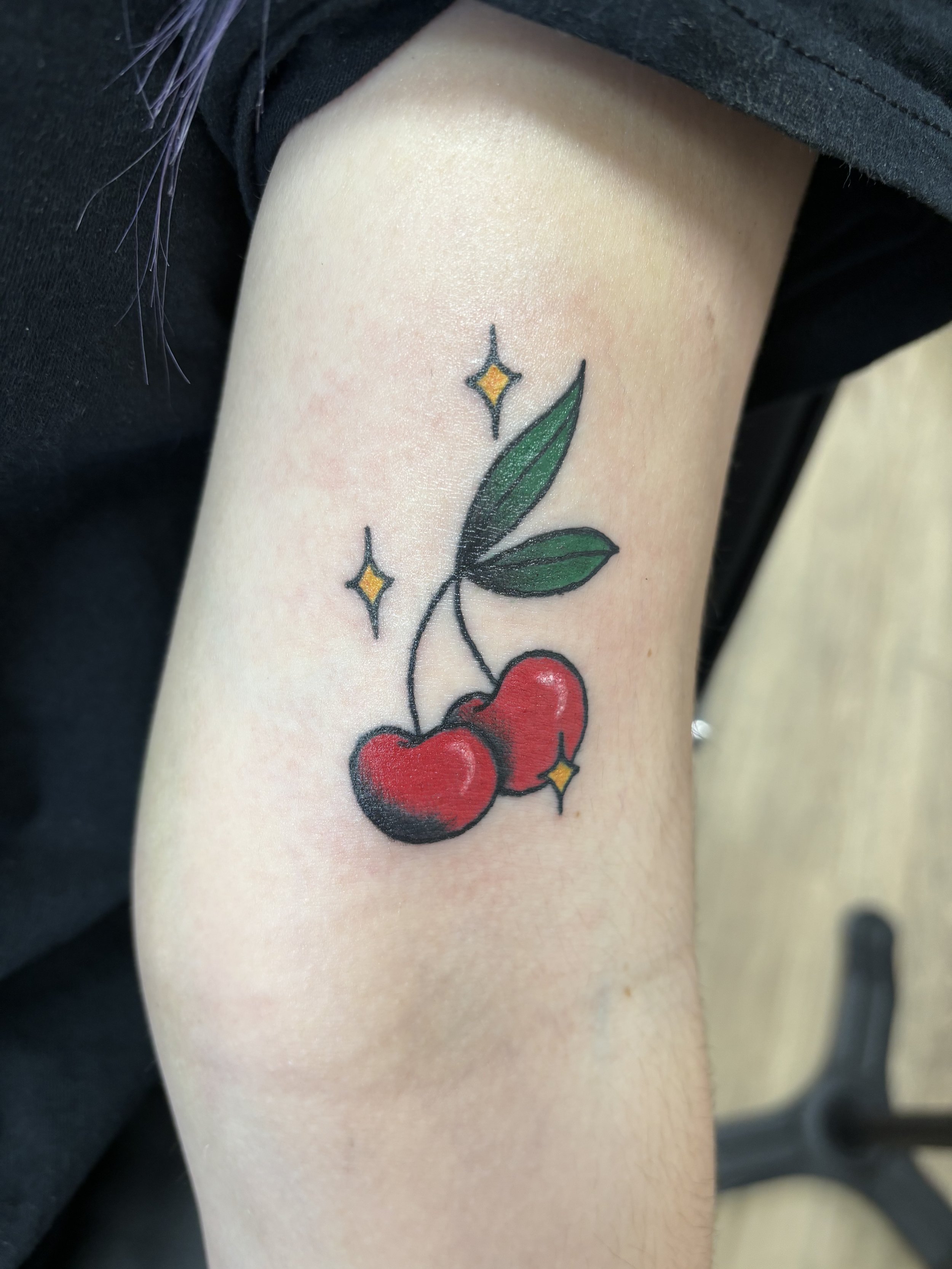 Tattoo of two red cherries with green leaves and yellow star accents on an arm.