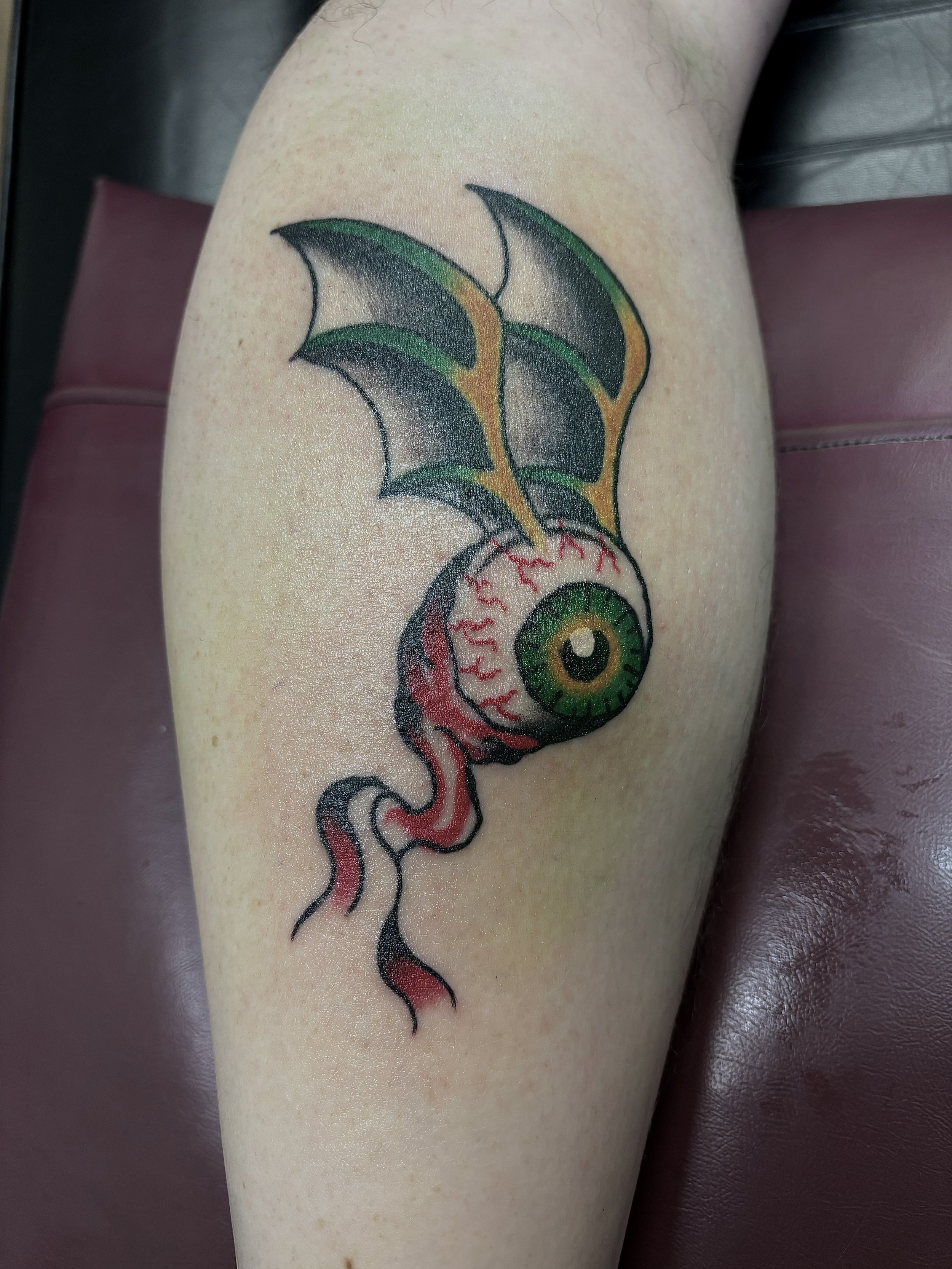 Tattoo of a green eyeball with bat-like wings and red tendrils on skin.