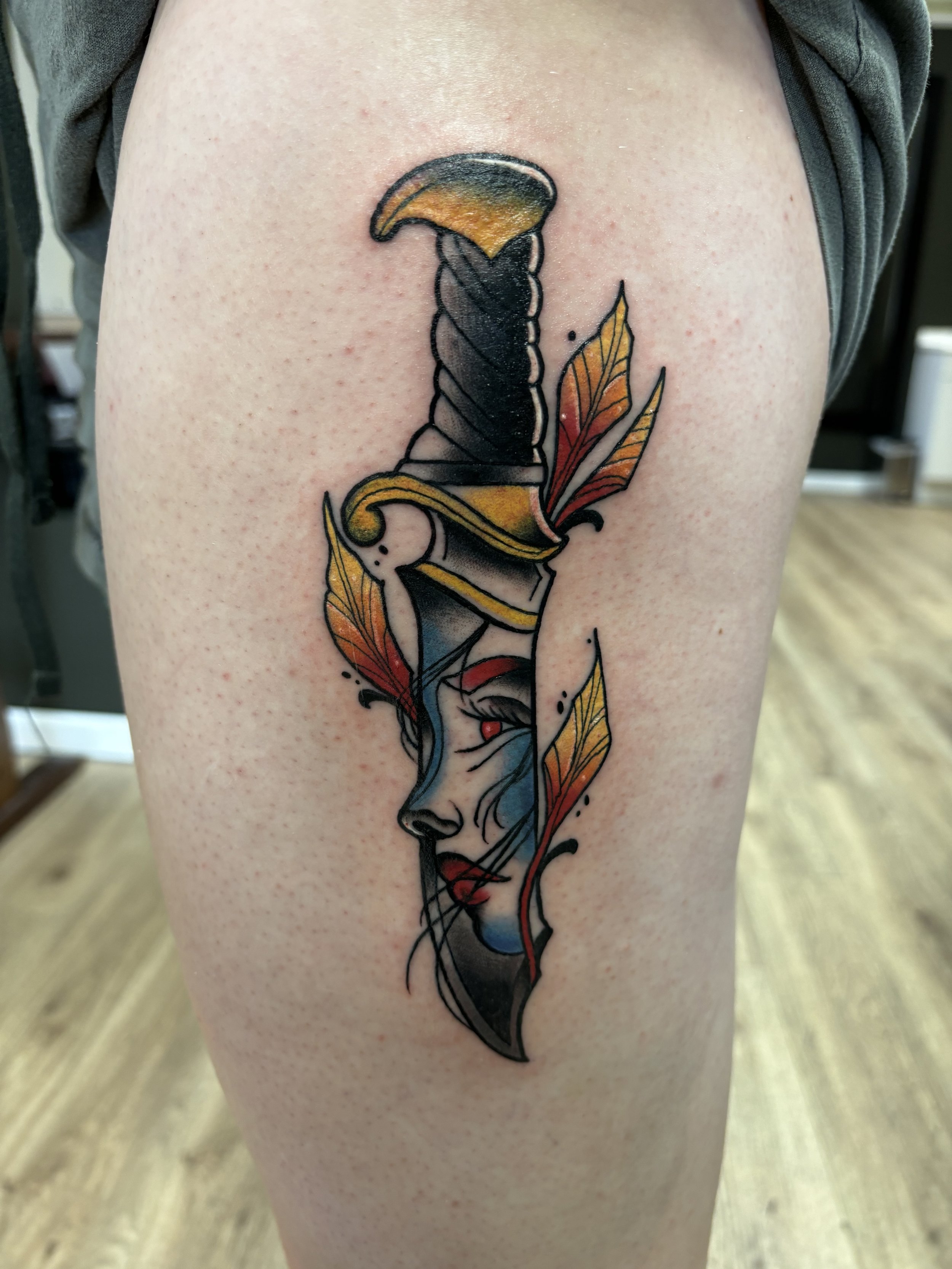Tattoo of a dagger with a woman's face in the blade, surrounded by leaves on skin.