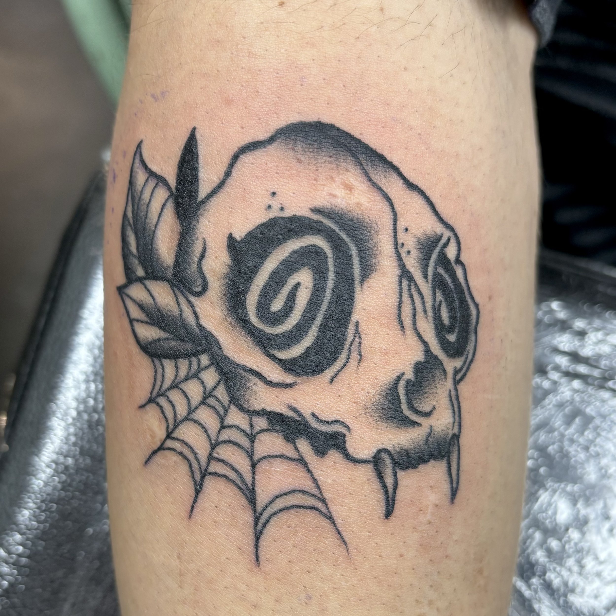Tattoo of a stylized skull with sharp fangs, spiral eyes, and a spiderweb pattern on the side.