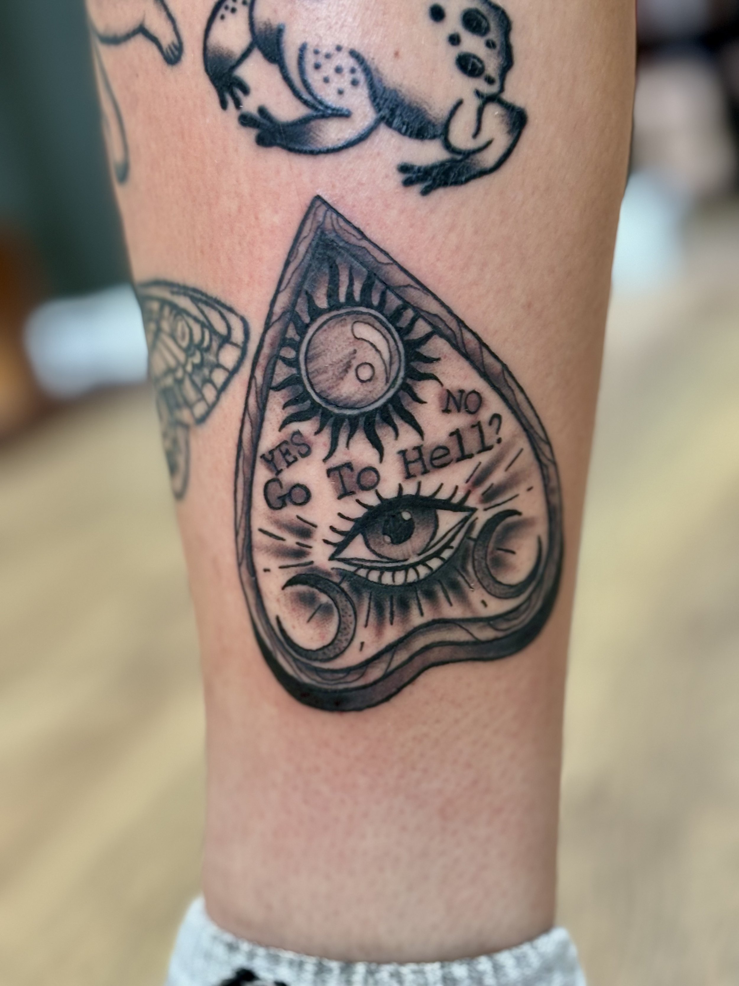 Tattoo of a Ouija board planchette with an eye design and the text 'Yes,' 'No,' and 'Go To Hell.'
