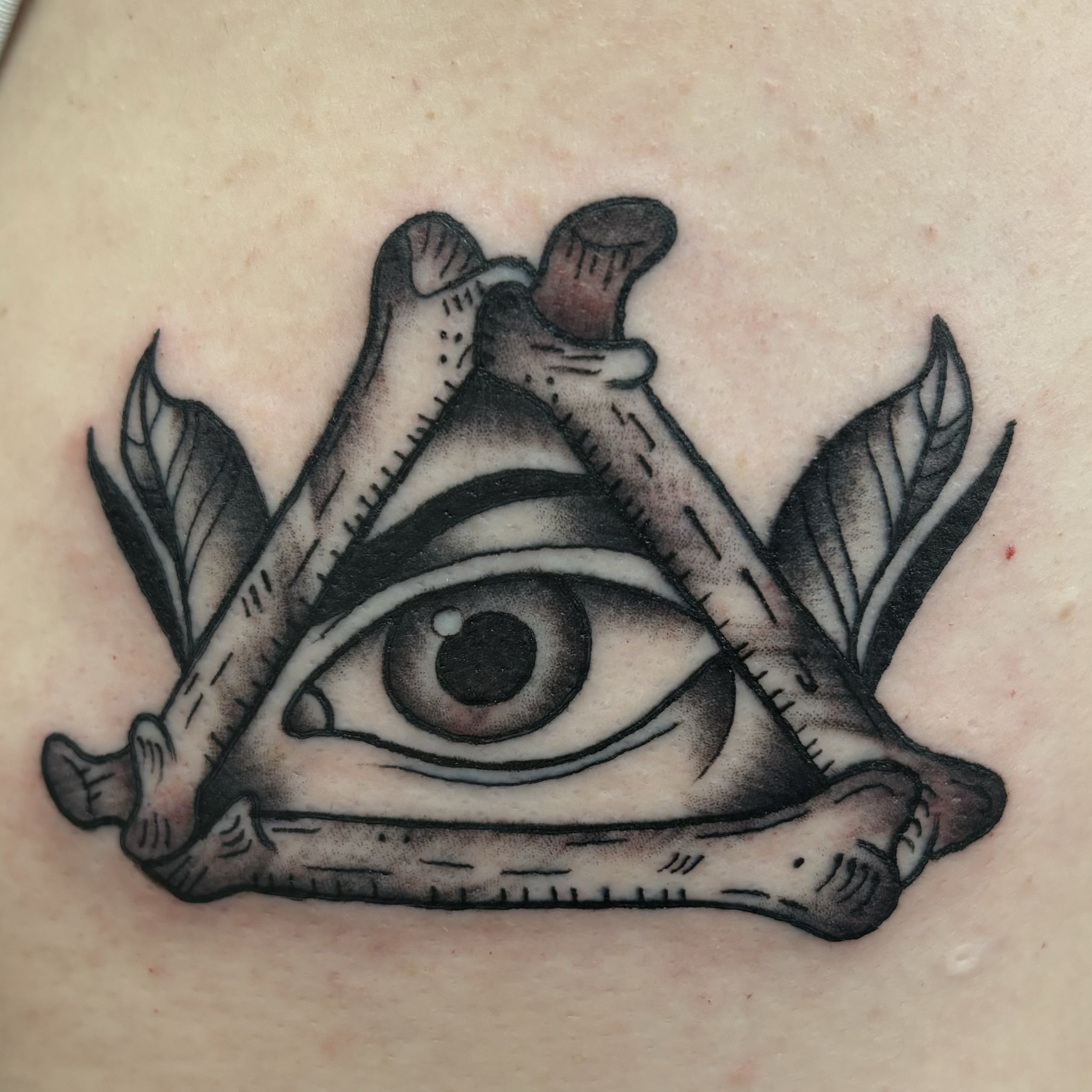 Tattoo of an eye within a triangle made of bones, with leaves on the sides.