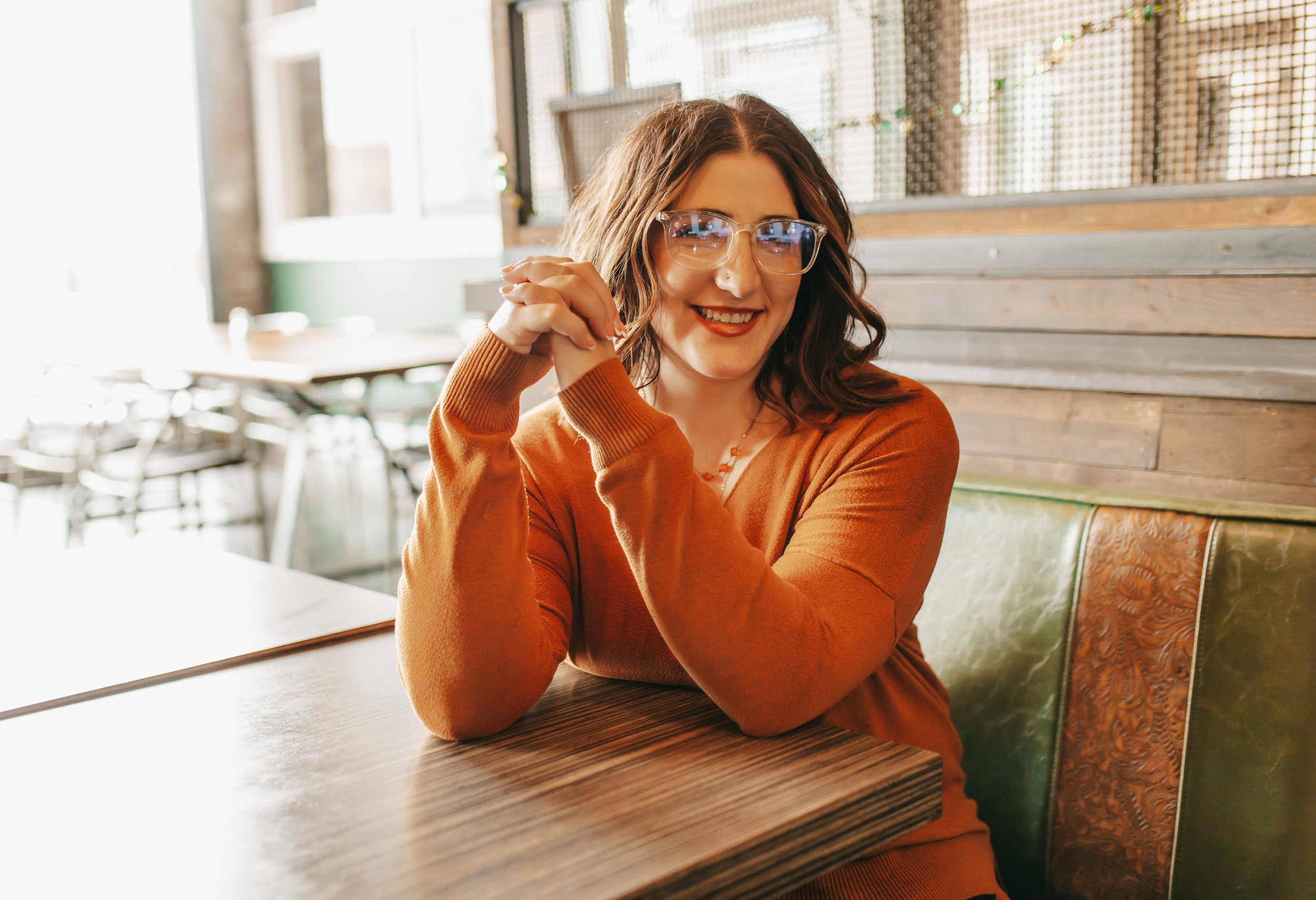 A woman with shoulder-length hair, wearing glasses and an orange sweater, smiling while sitting at a wooden table in a cozy cafe with wood paneling and string lights in the background.