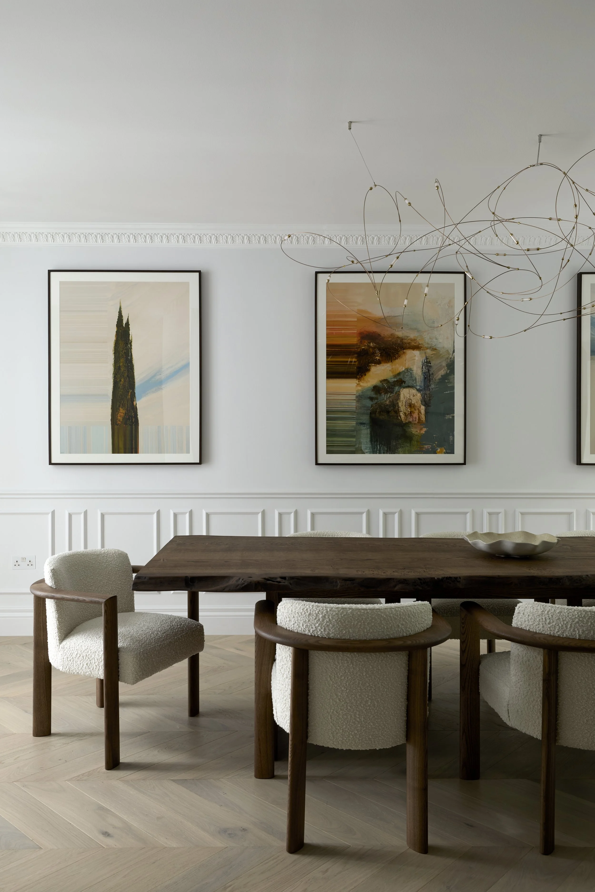 A dining room with four upholstered chairs around a dark wooden table, four abstract landscape paintings on the wall, and a modern light fixture hanging from the ceiling.