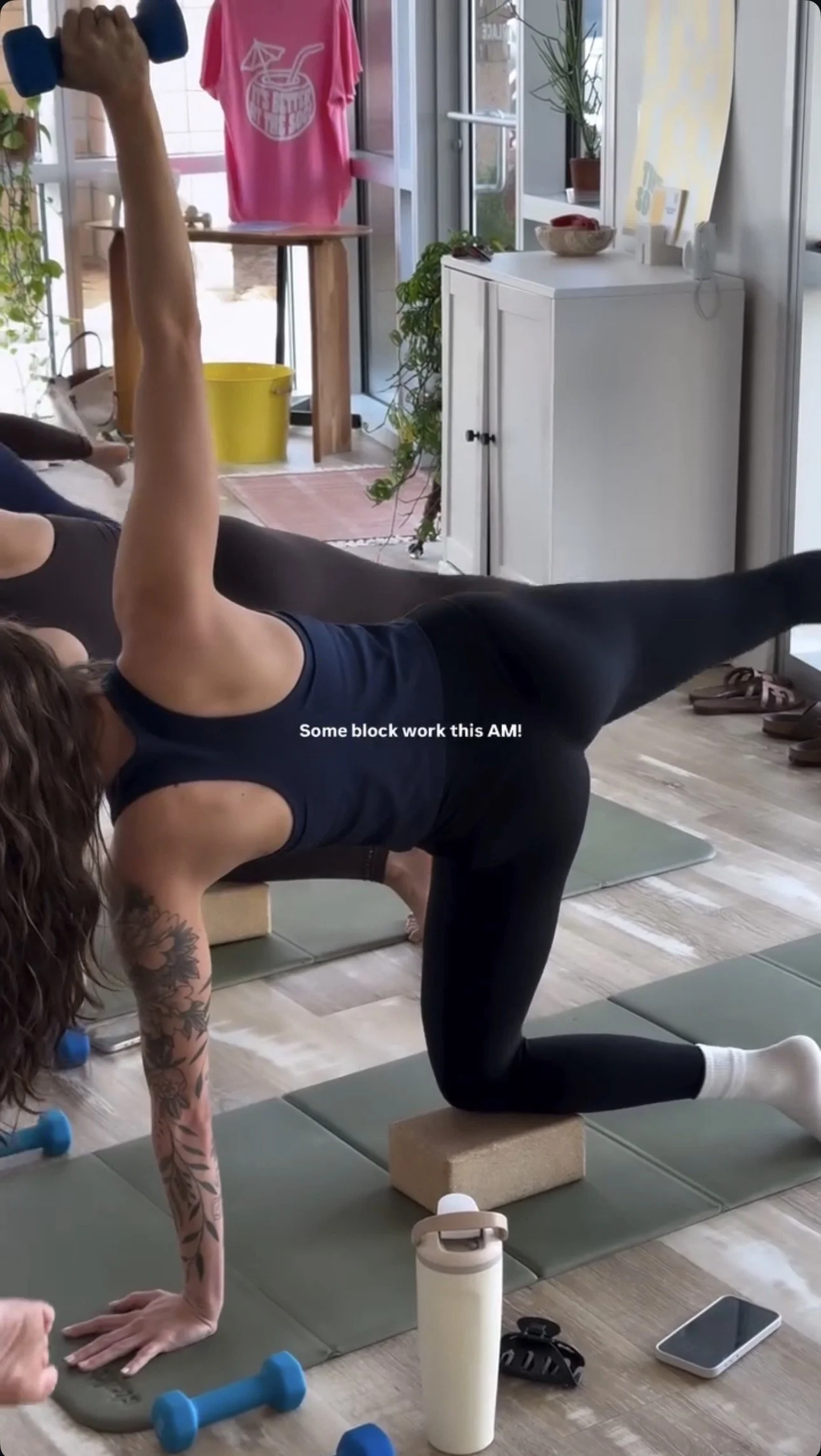 A woman taking a selfie in a fitness studio, wearing a black Peloton sports bra and pink leggings. She is smiling, holding a green resistance band labeled "PAGE F" with her right hand. There are yoga mats on the wooden floor, large windows in the background, and a few indoor plants. Text overlay reads "I broke a band @the_society_wellness" and "ROCK YOUR BODY".
