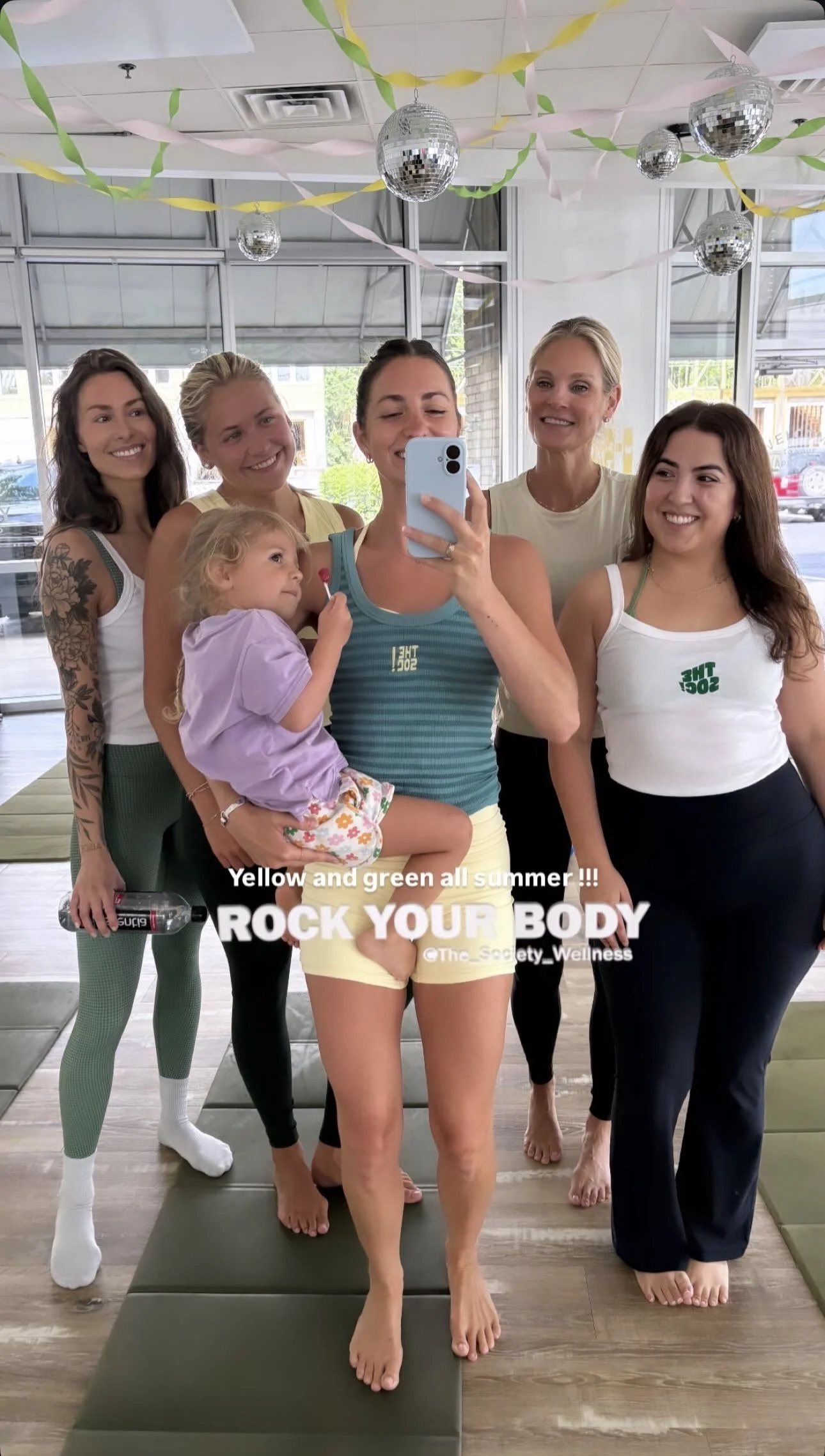 Two women in athletic wear taking a mirror selfie at a wellness or fitness center. One woman is wearing a pink sports bra and matching leggings, and the other is wearing a blue sports bra and black shorts. They are barefoot, standing on yoga mats, and making peace signs. The background shows a modern, well-lit interior with large windows and fitness equipment. Text on the image reads 'ROCK YOUR BODY' and '@the_society_wellness'.