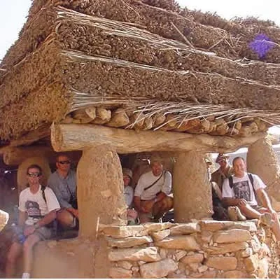 Drive The Globe expedition team sitting under a large, traditional mud and straw structure supported by stone pillars, likely at an archaeological site.