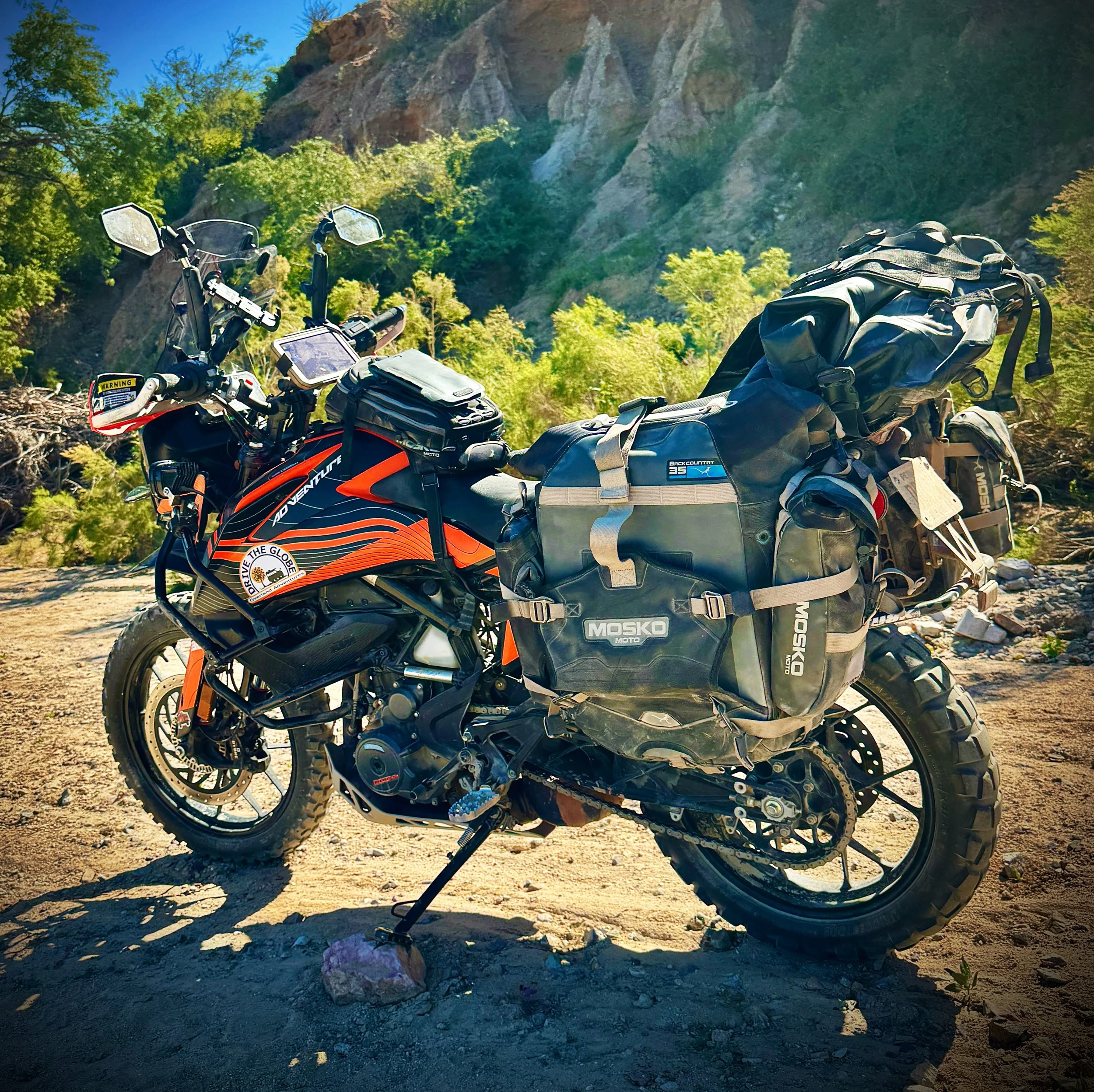 A KTM 390 adventure motorcycle equipped with large Mosko Moto saddlebags and gear is parked on a dirt surface in a mountainous area with green trees and rocky cliffs in the background.