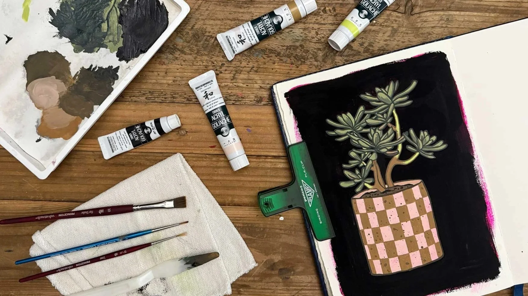 Painting of a succulent in a sketchbook laying flat on a desk covered in art supplies.