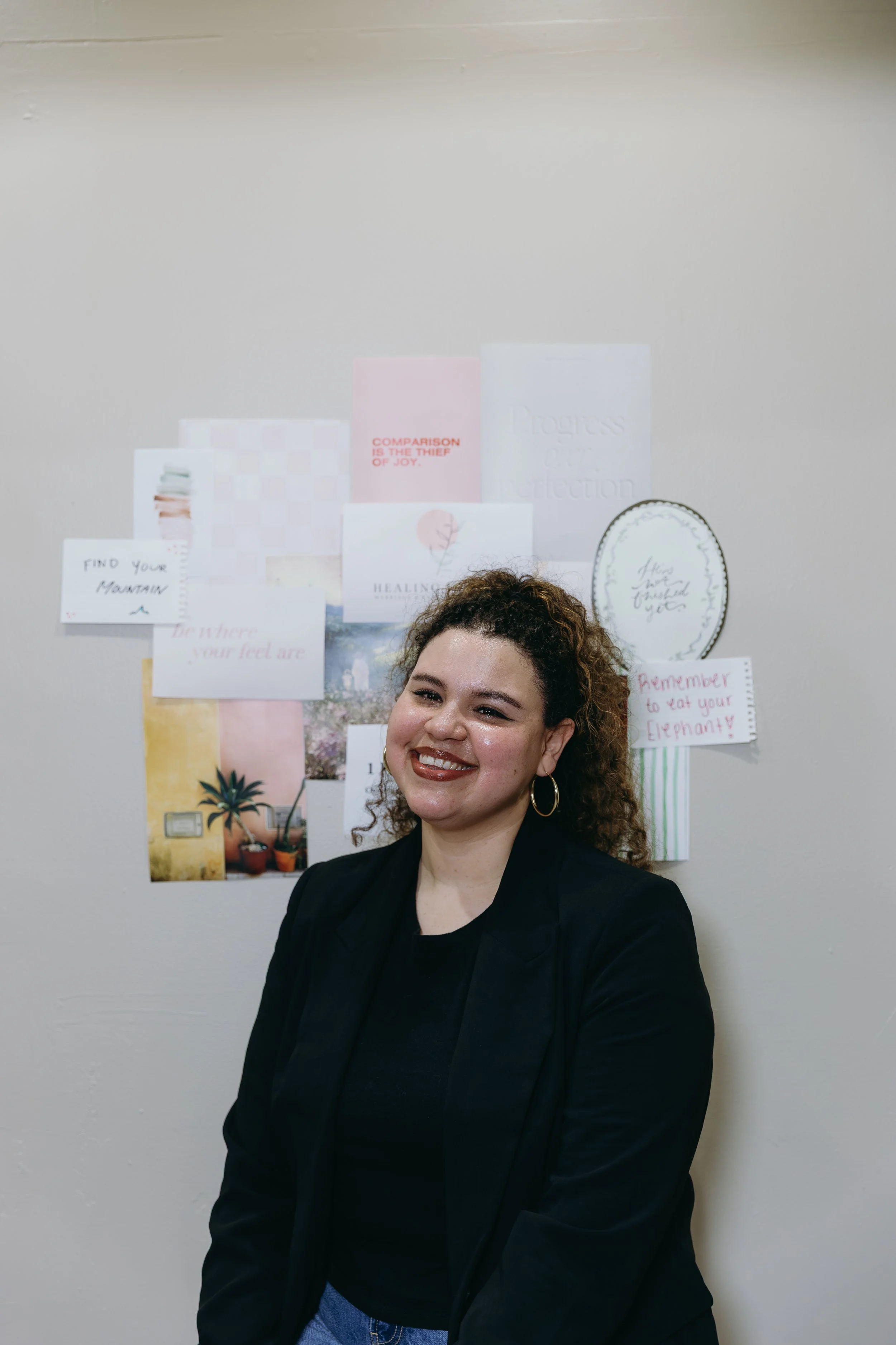 A young woman with curly hair and hoop earrings smiling while sitting on a park bench outdoors, with houses and greenery in the background.