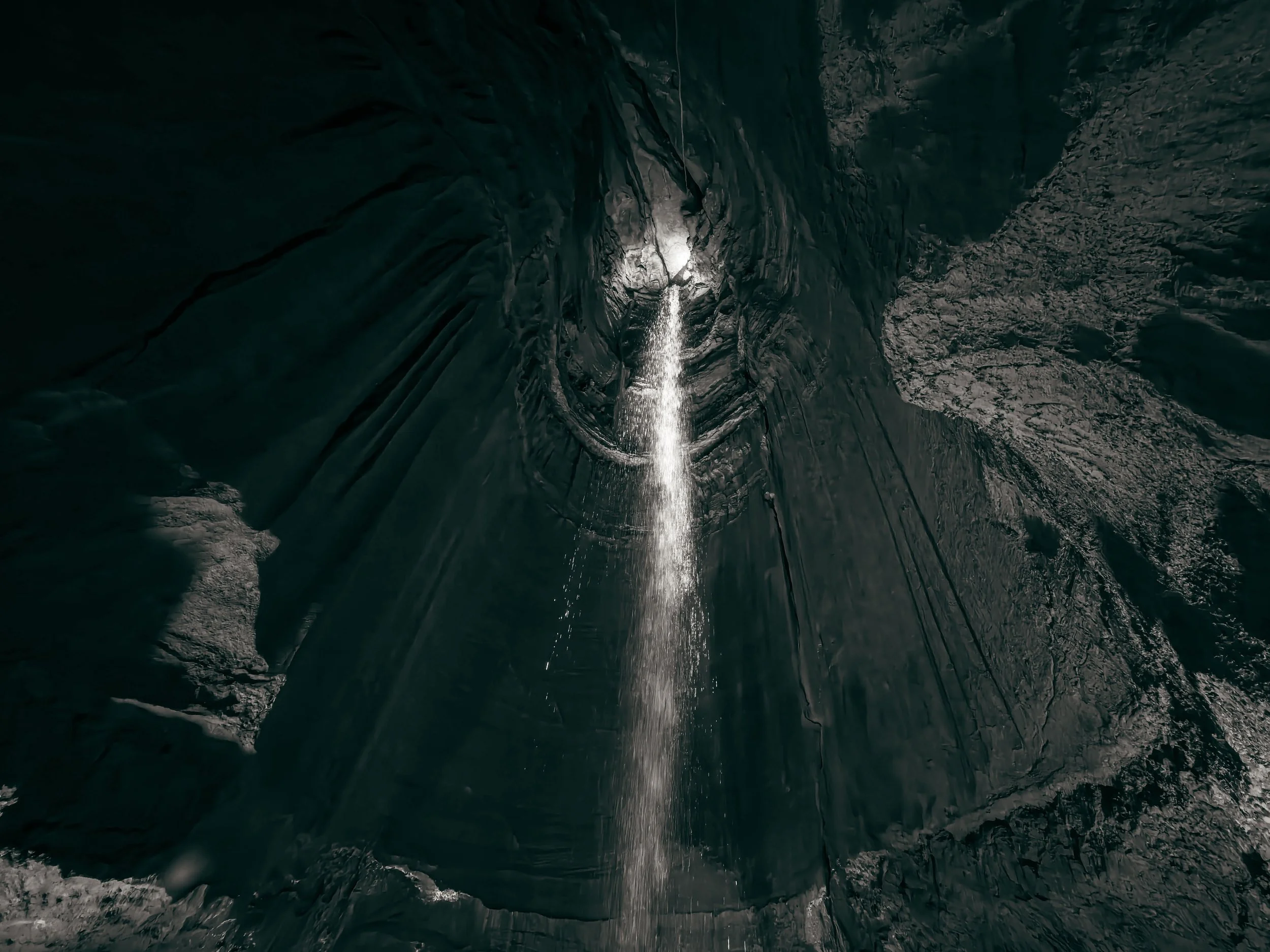 View of a dark rocky cave with a small waterfall of water falling from above. Light illuminates part of the water and rocks inside the cave.