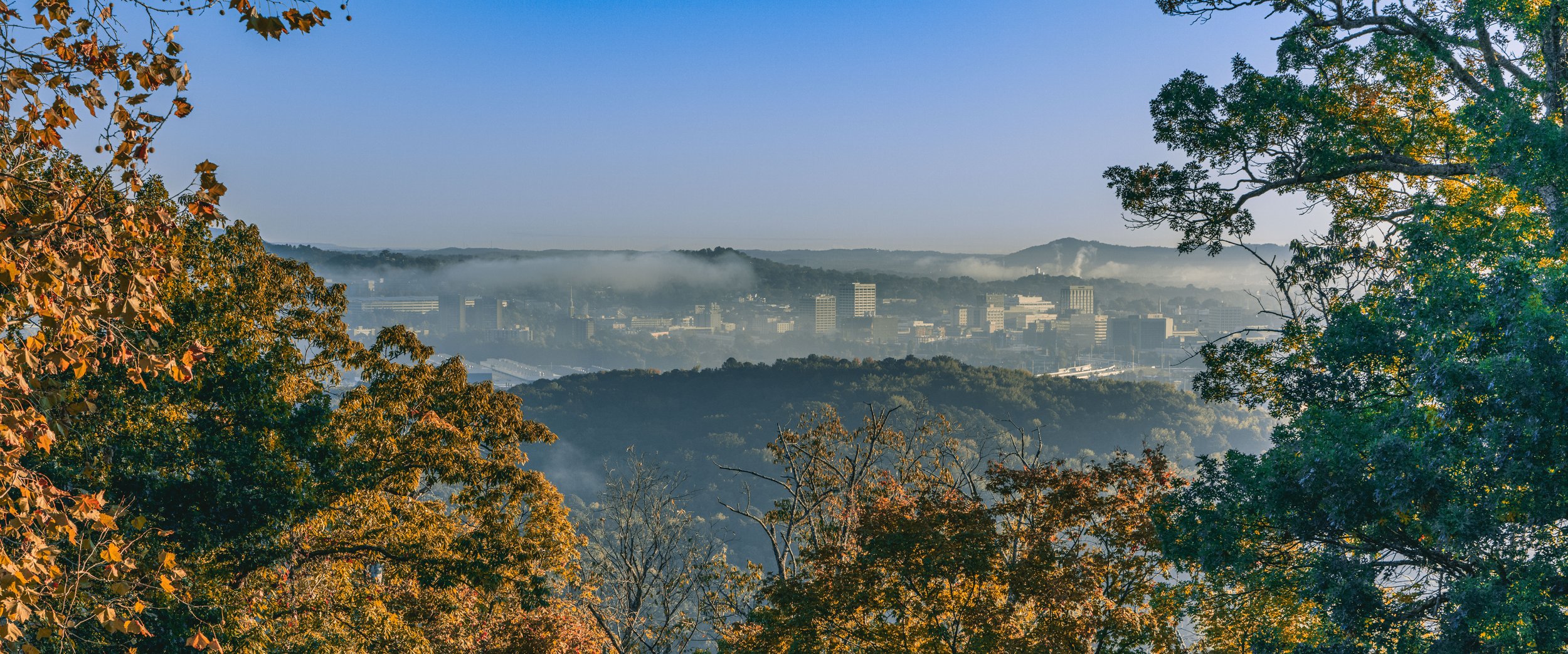 A cityscape seen through a frame of trees with autumn-colored leaves, fog rising over buildings, and hills in the background.