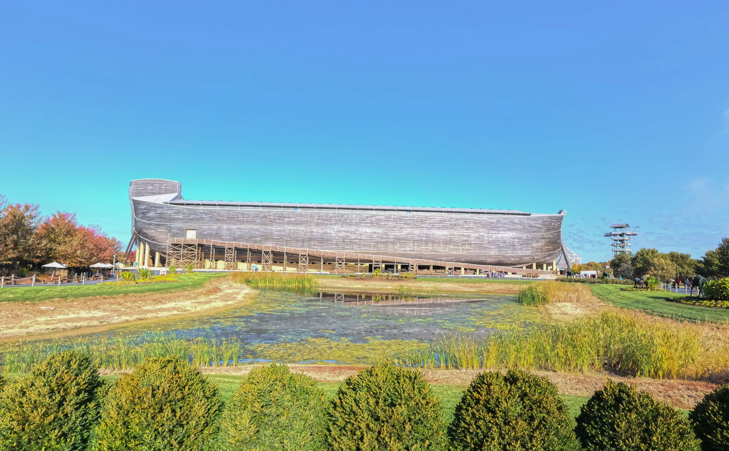 The image shows a large wooden ship-shaped building in a park-like setting with trees, a pond, and a clear blue sky.