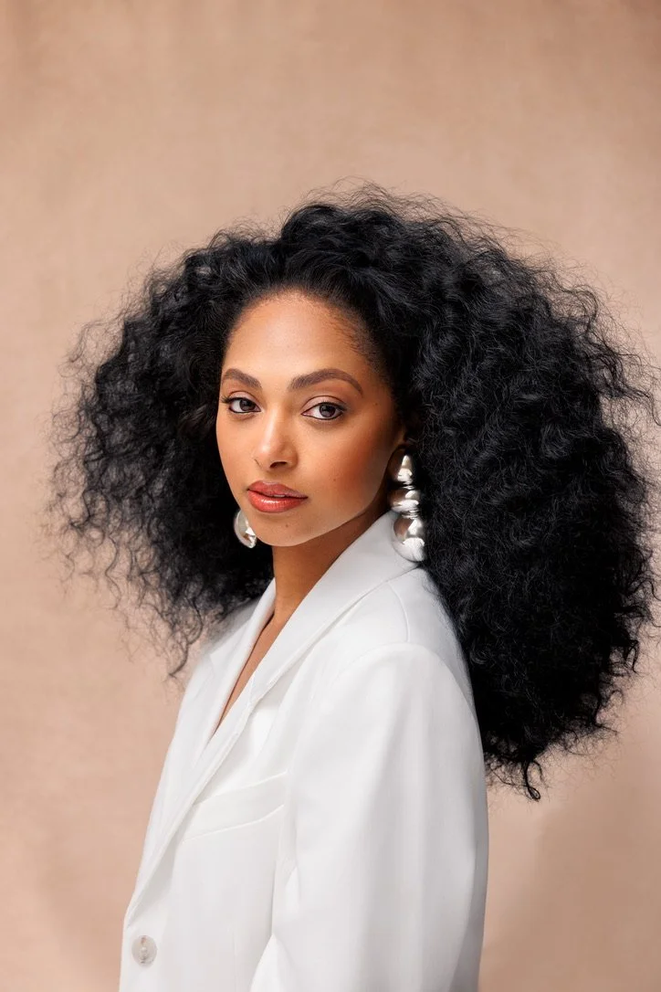 A woman with dark, voluminous, curly hair wearing a white blazer and large silver earrings, looking directly at the camera against a beige background.