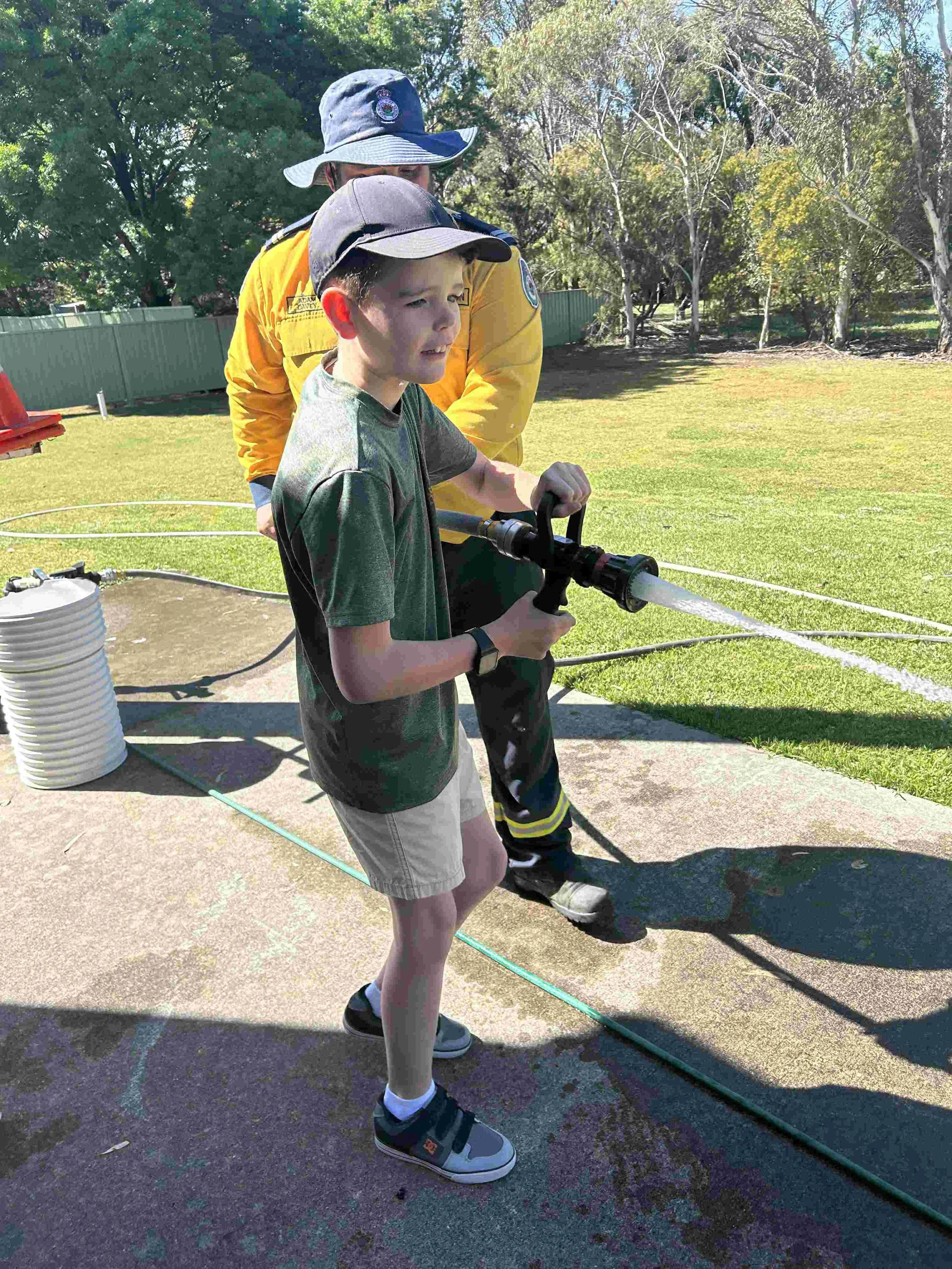 A young boy is holding a garden hose, spraying water with the guidance of a firefighter in yellow protective gear, outdoors on a sunny day with trees and a green fence in the background in Bathurst NSW