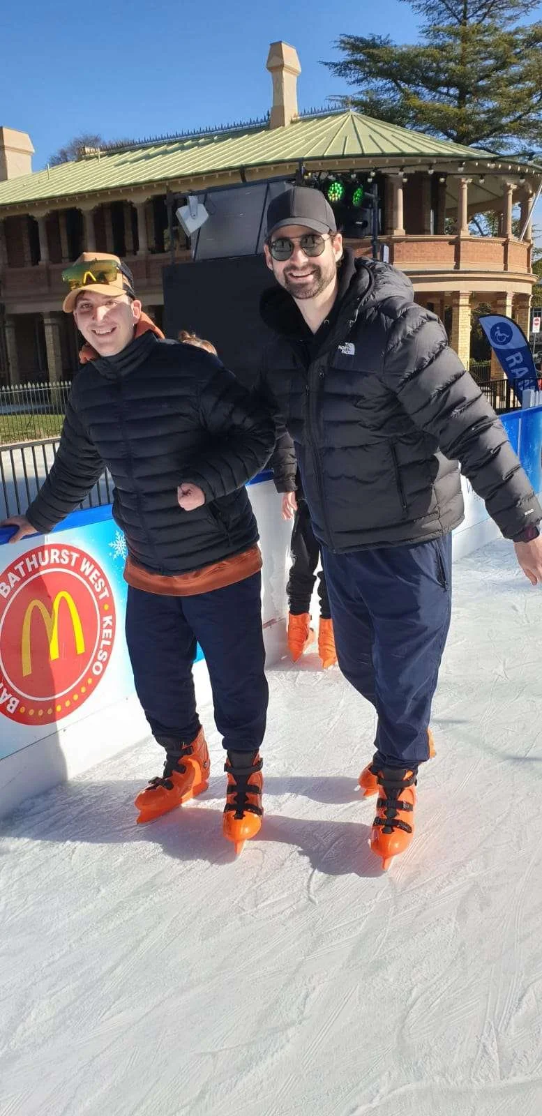 Two men ice skating outdoors, wearing black jackets and orange ice skates, smiling at the camera, with a historic building and a sign with the McDonald's logo in the background.