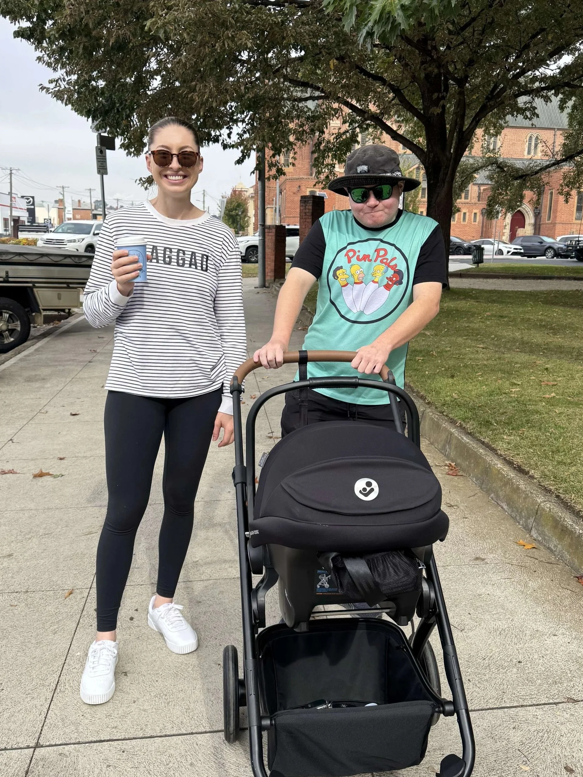 A woman and a man with a stroller in a park. The woman is smiling, wearing sunglasses, a striped shirt with 'BAD GAD' written on it, black leggings, and white sneakers. The man is wearing dark sunglasses, a black and teal T-shirt with a cartoon logo, a bucket hat, and dark shorts.