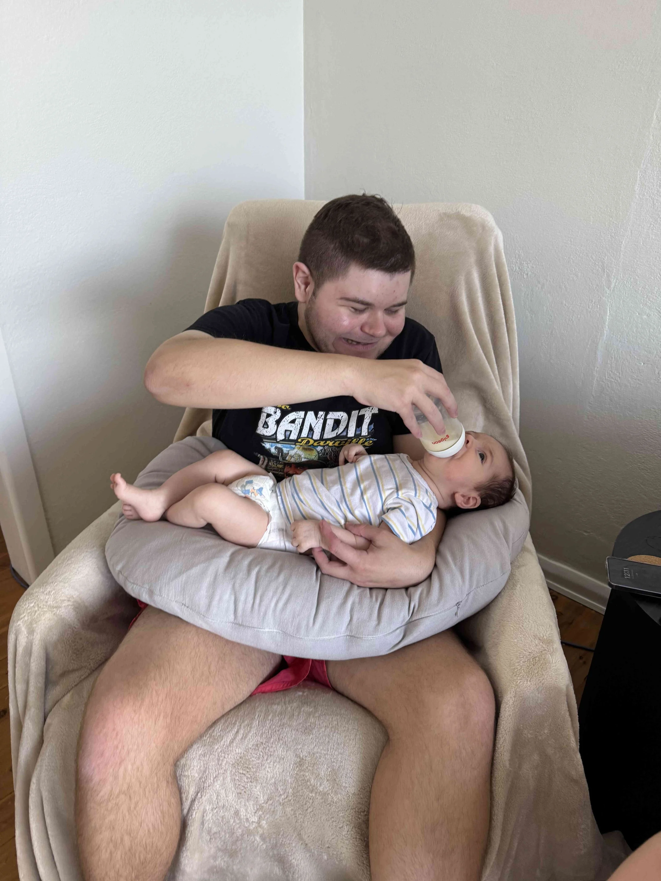 A young man sitting in a beige armchair, feeding a baby bottle to a baby lying on a nursing pillow on his lap.