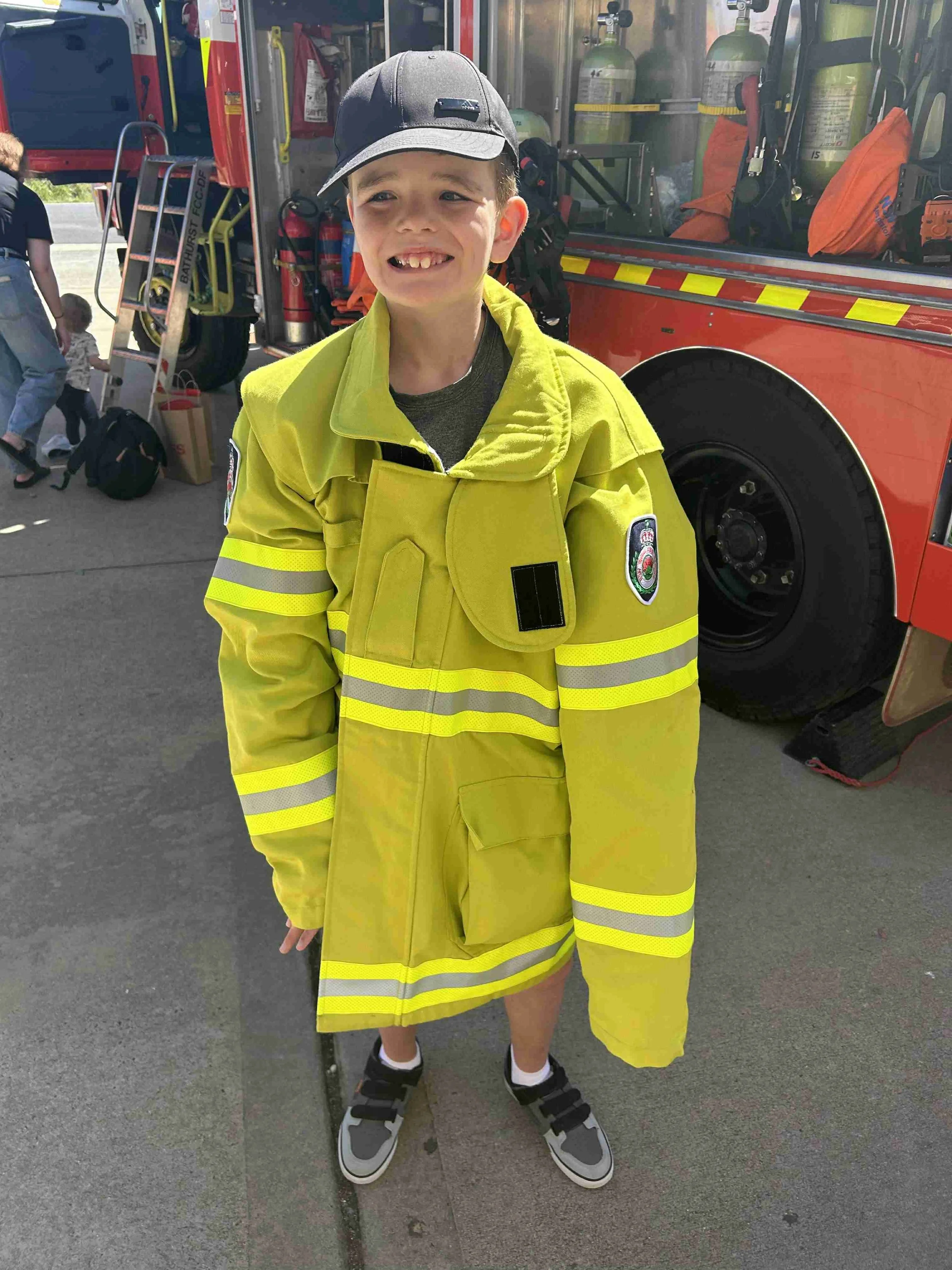 Young boy smiling, dressed in firefighter gear, standing in front of a fire truck.