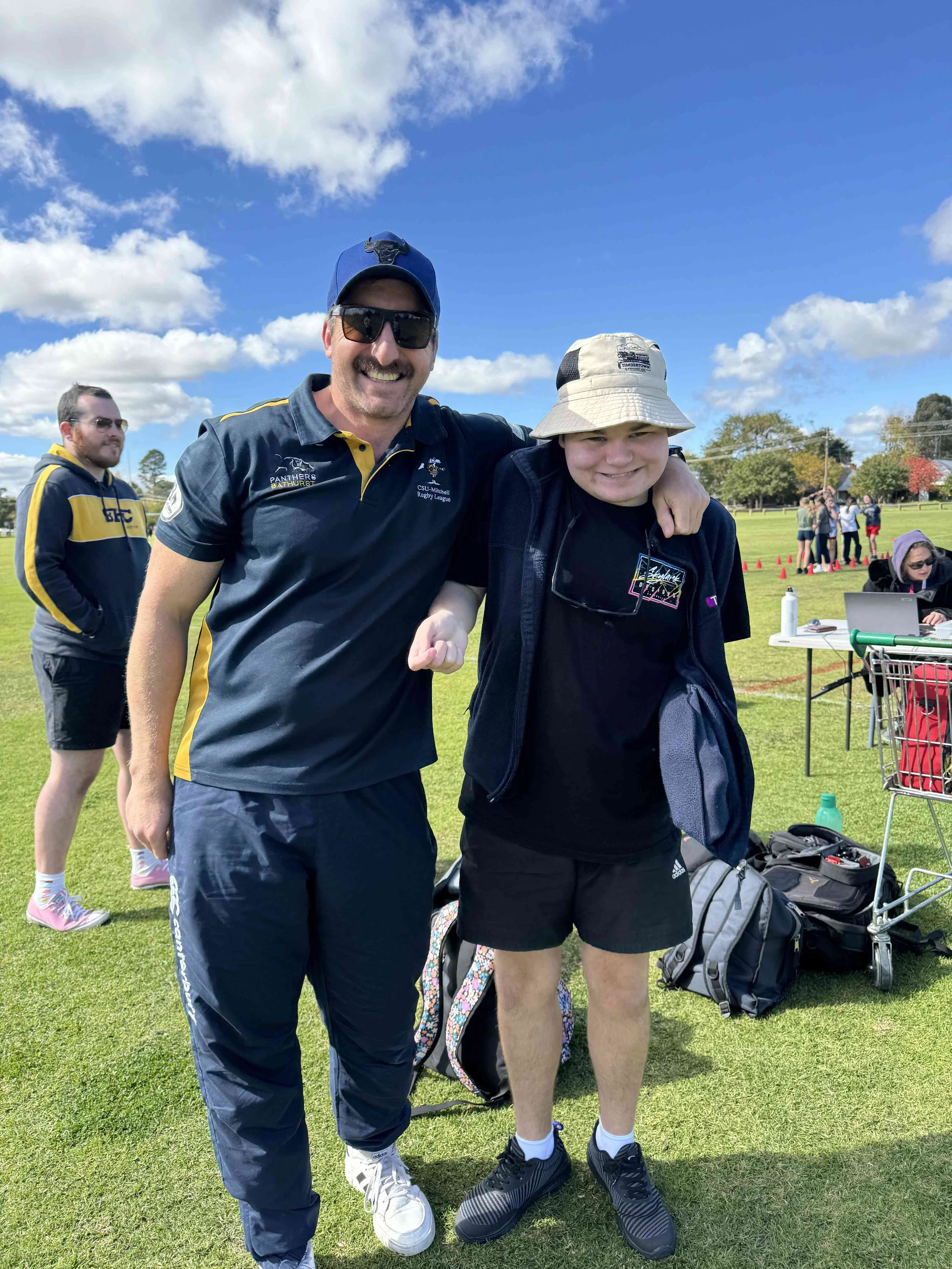 Two people smiling and posing for a photo outdoors on a grassy field on a sunny day. The person on the left is wearing sunglasses, a navy blue baseball cap, a navy blue sports shirt with yellow accents, and navy blue pants. The person on the right is wearing a bucket hat, glasses hanging on a shirt, black shorts, and black sneakers. In the background, there are other people, some at a table with a laptop and bags, and a group of people near orange cones