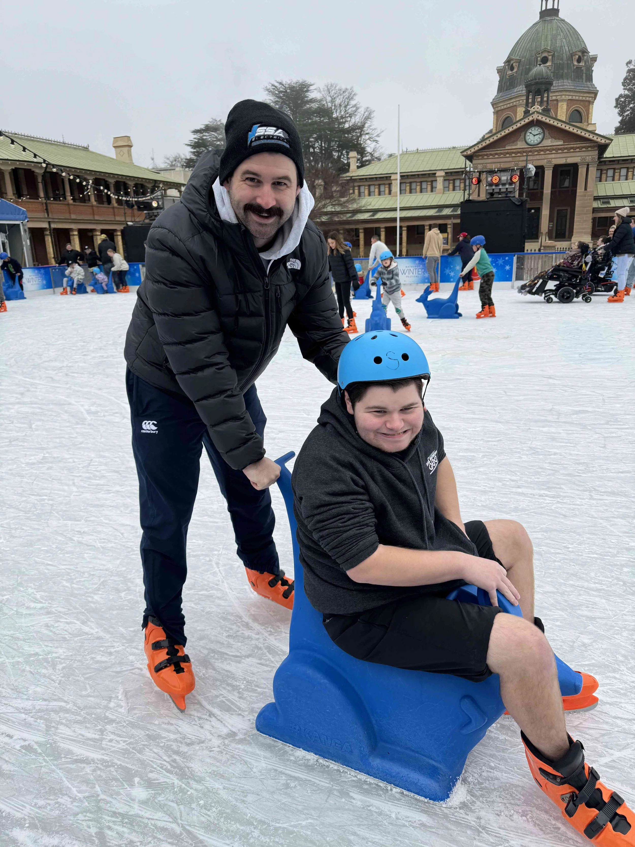 A man and a young boy at an outdoor ice skating rink, with the man pushing the boy on a blue penguin-shaped skating aid, both wearing helmets and skates, surrounded by other skaters and a historic building in the background.