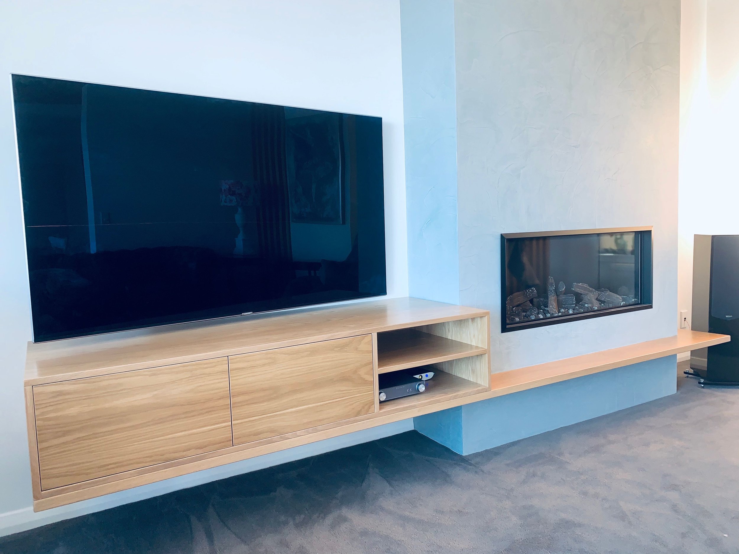 American white oak entertainment unit consiting of shelves and cabinets, a wall hung tv hangs above and a fireplace to the right with a oak shelf below