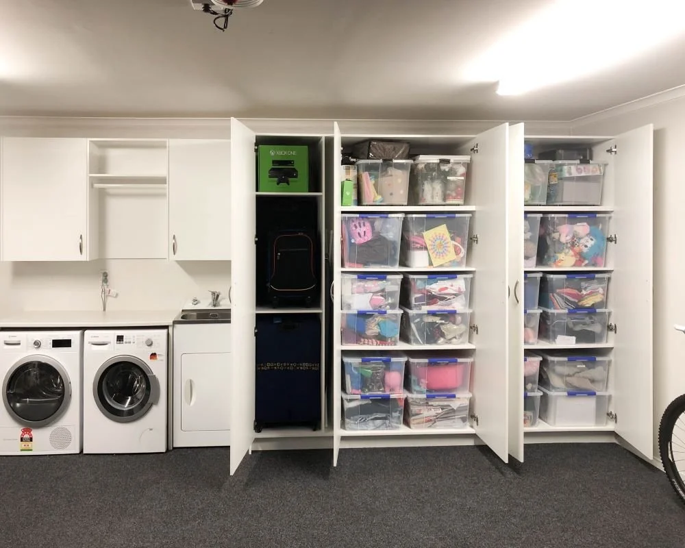 White melteca tall and wall hung cabinets built around washing sink, washing machine and dryer which are visible. Tall cabinet units doors are open and storage containers are stacked on shelves 