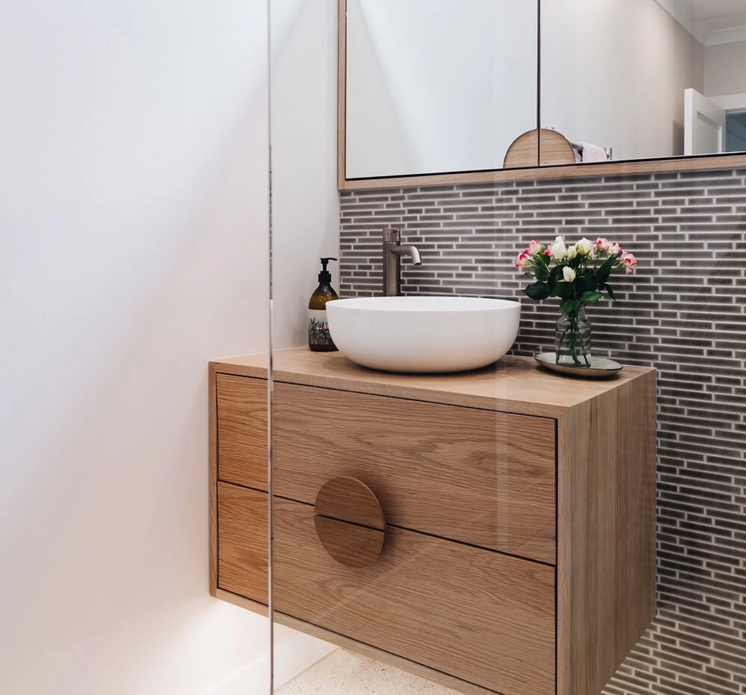 Timber look two drawer vanity with white sink top and chrome tapware. A vase with pink tipped flowers sits to the right on vanity top and a pump handsoap to the left. A wall hung mirror hangs above vanity with long thin times on the wall