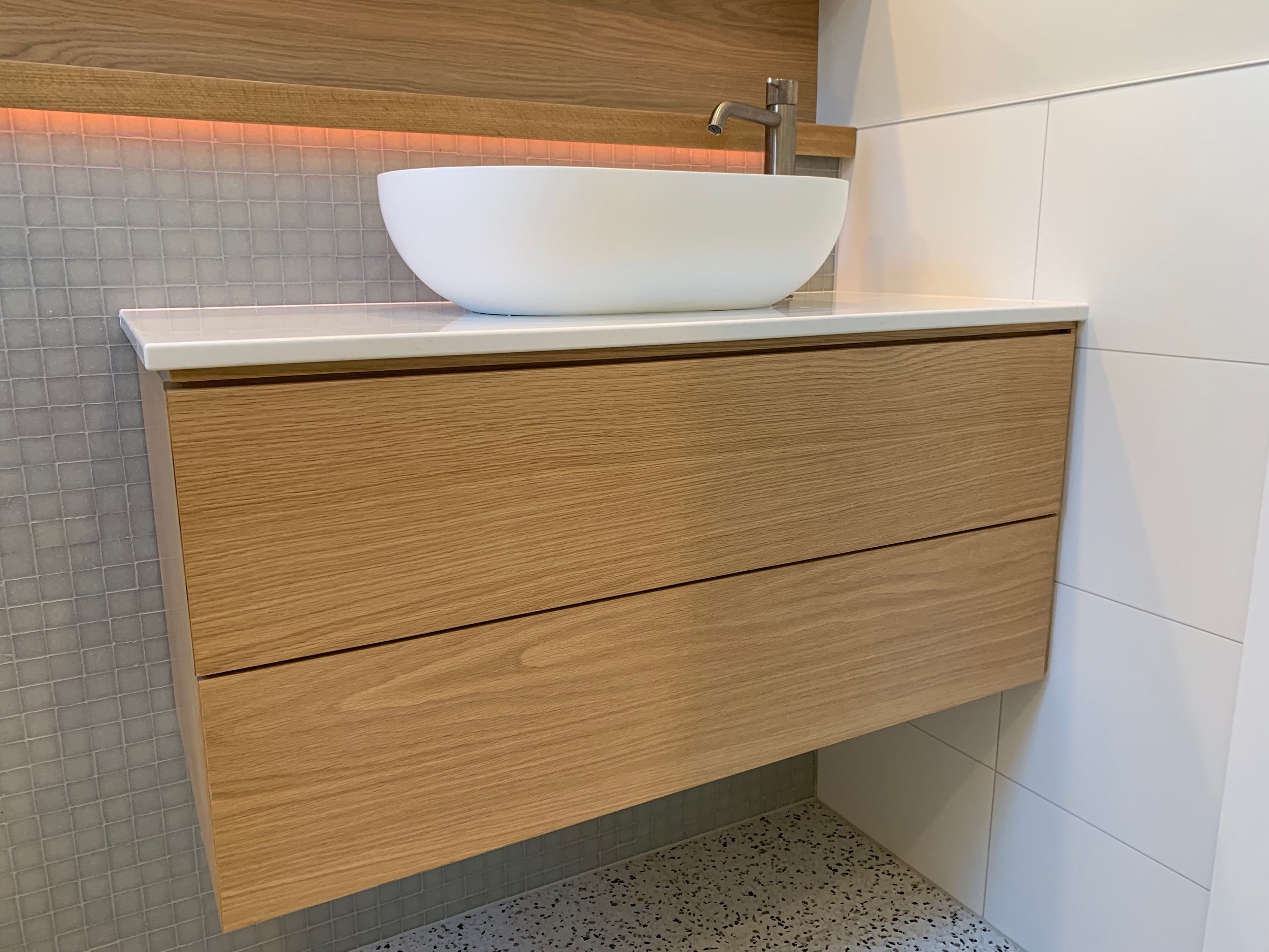 Timer look two drawer vanity with white bench top and white basin. Chrome tapware over basin. Small beige tiles behind basin. and large white rectangle tiles to the left