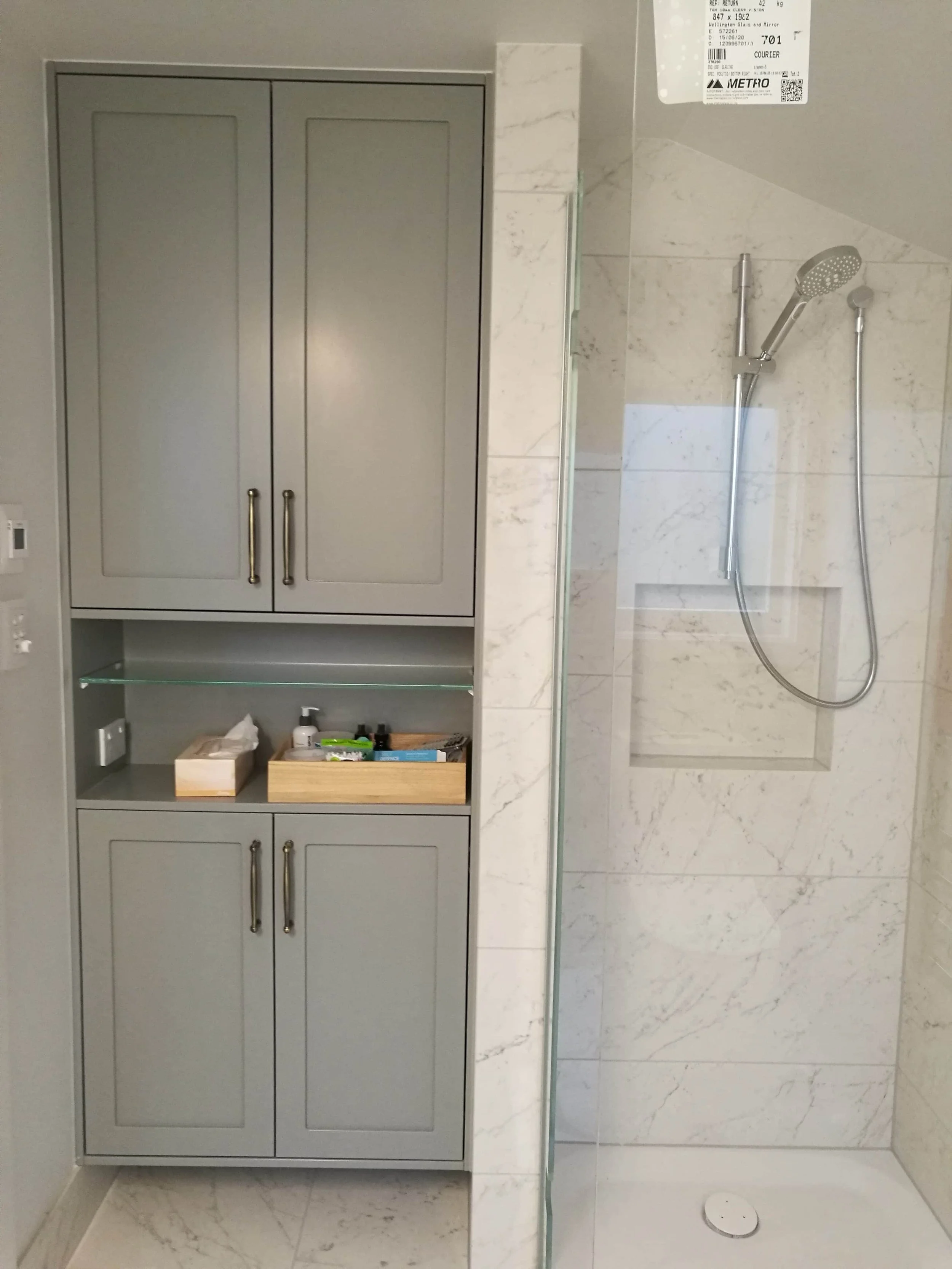 Built in grey bathroom storage cabinets. Double door cabinet at top and bottom with a shelf in the middle with oak box containing bathroom products viable and box of tissues. Shower with chrome tapware and glass door visible to the left