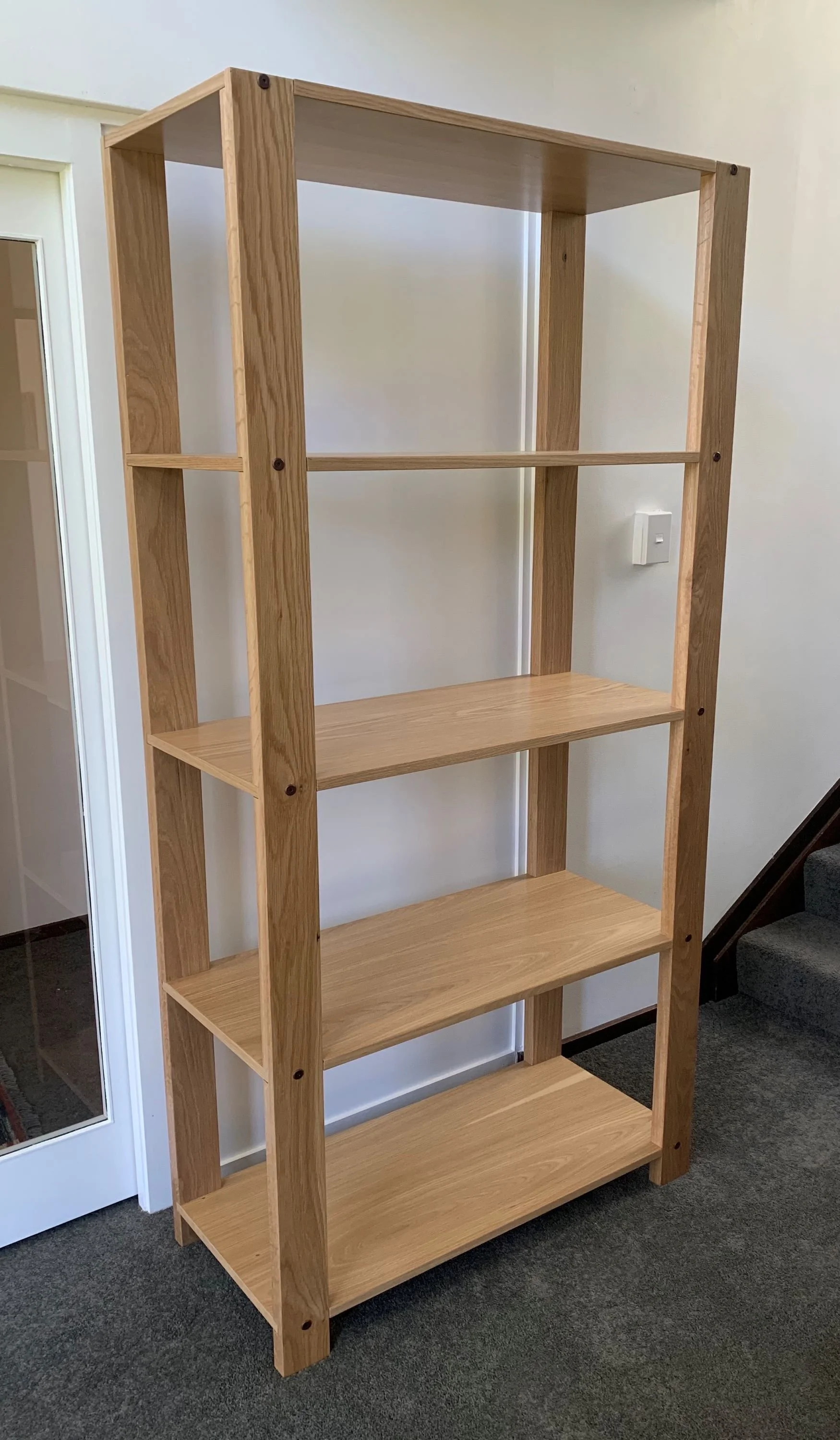American white Oak shelving unit with five shelves up against white wall with carpeted stairs to the right