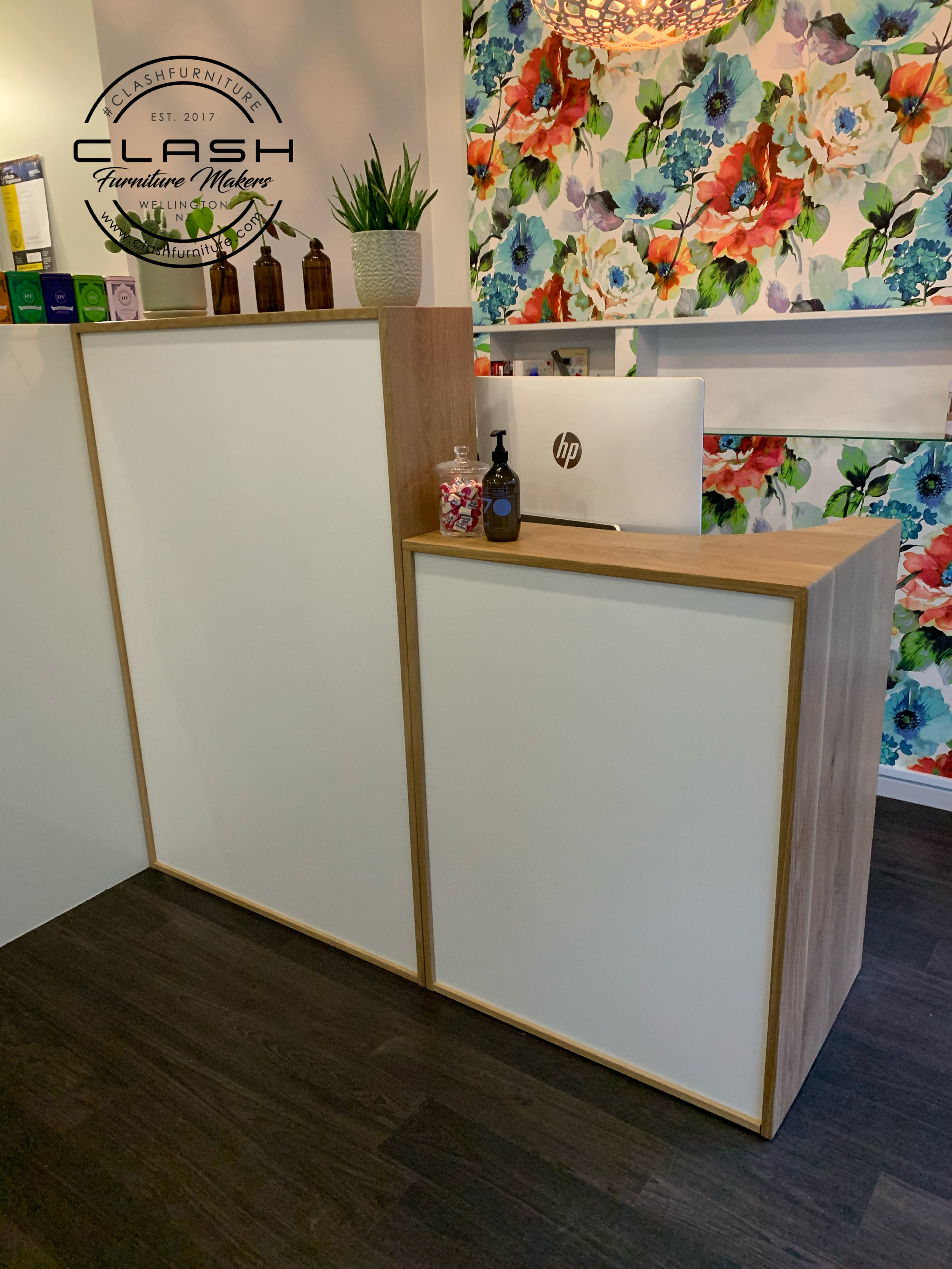 White melteca and oak reception desk with colourful flower wall paper in background. An back of an HP computer is visible on the desk as well as product bottles and a jar of lollies